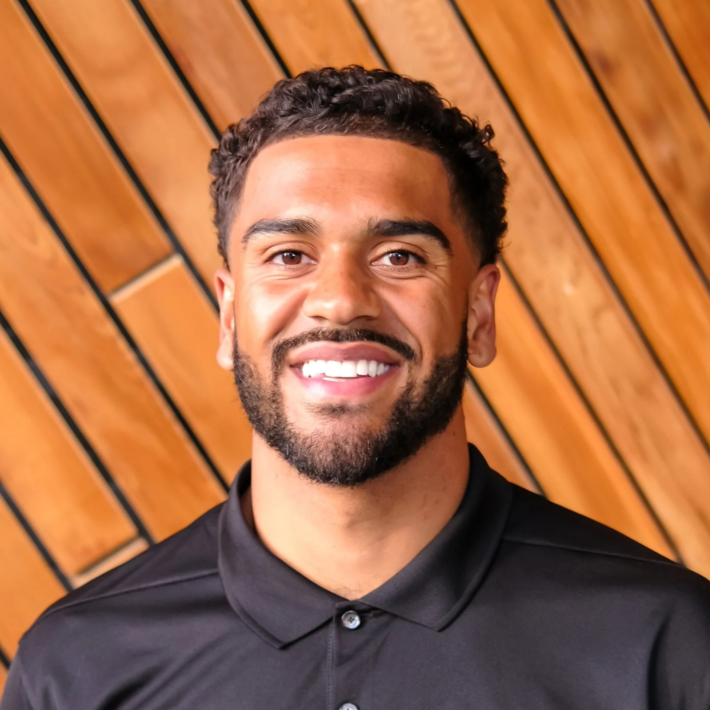 A smiling young man with dark curly hair and a beard wearing a black collared shirt, standing against a wooden wall background.