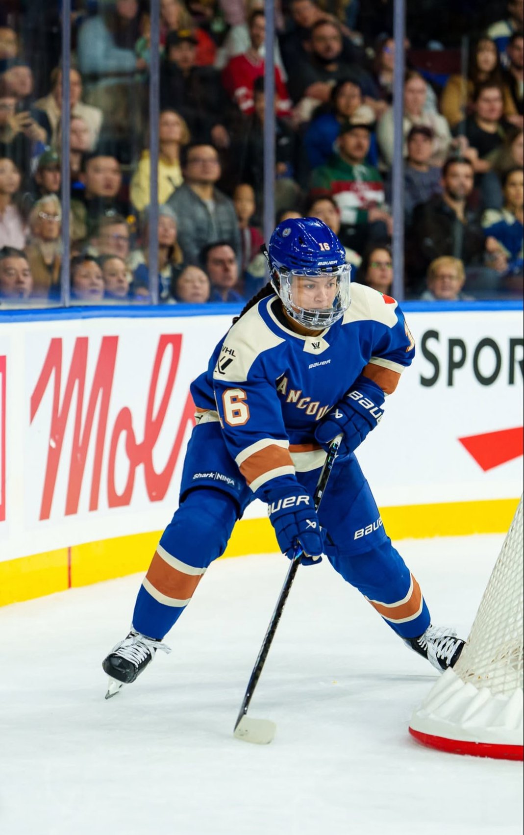 Hockey player in blue and white uniform with orange accents, numbered 16, skating on ice near the goal, holding a hockey stick, with a crowd of spectators in the background.