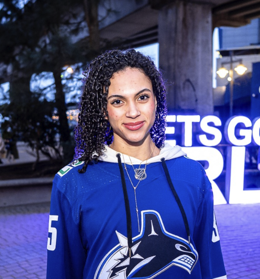 Young woman with dark, curly hair smiling in a blue San Jose Sharks hockey jersey, with a neon sign in the background.