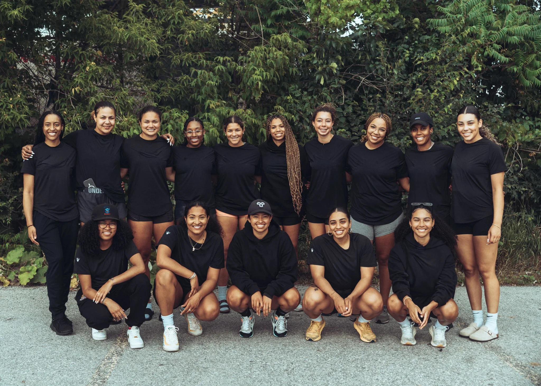 A group of fifteen women wearing black athletic shirts, posing outdoors in front of green bushes, with some standing and others squatting, smiling for the camera.