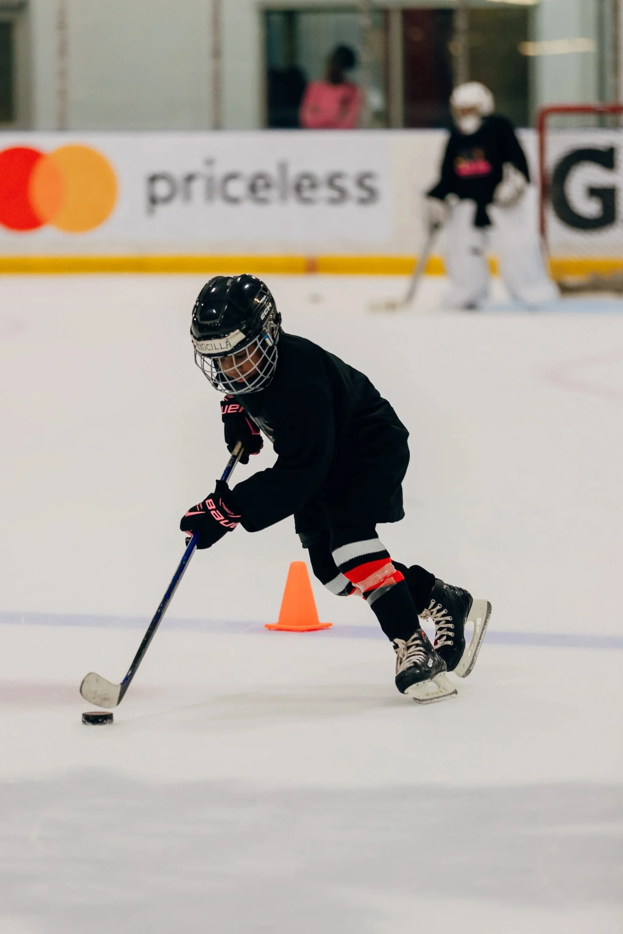Young hockey player wearing a black jersey, helmet, and gloves, skating on ice and controlling the puck with a stick, with an orange cone in front and an rink in the background, including a goalie and advertising banners.