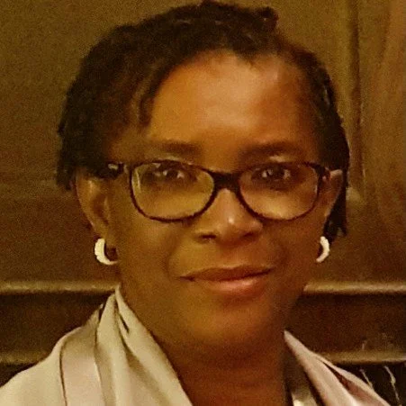Close-up of a woman with short hair, wearing glasses, earrings, and a white scarf, smiling in an indoor setting.