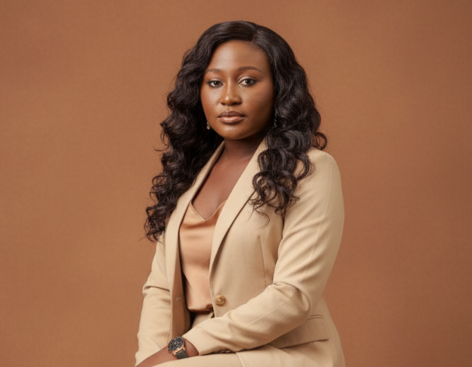 A woman with curly black hair wearing red glasses, a pearl necklace, and a black top. She is smiling and wearing a blue lanyard.