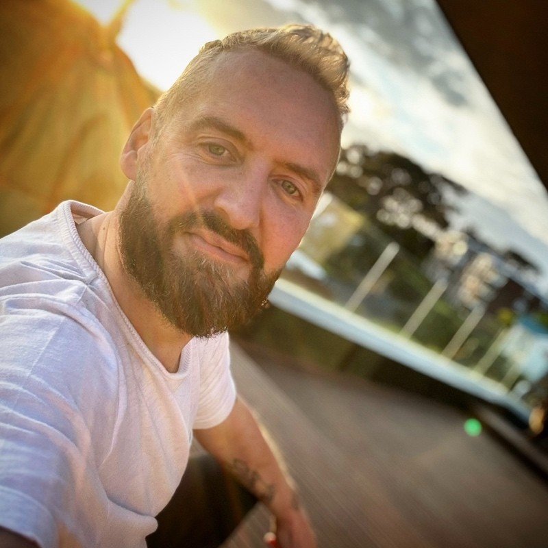 Close-up of a man with a beard and short hair, smiling outdoors during sunset, wearing a white T-shirt.