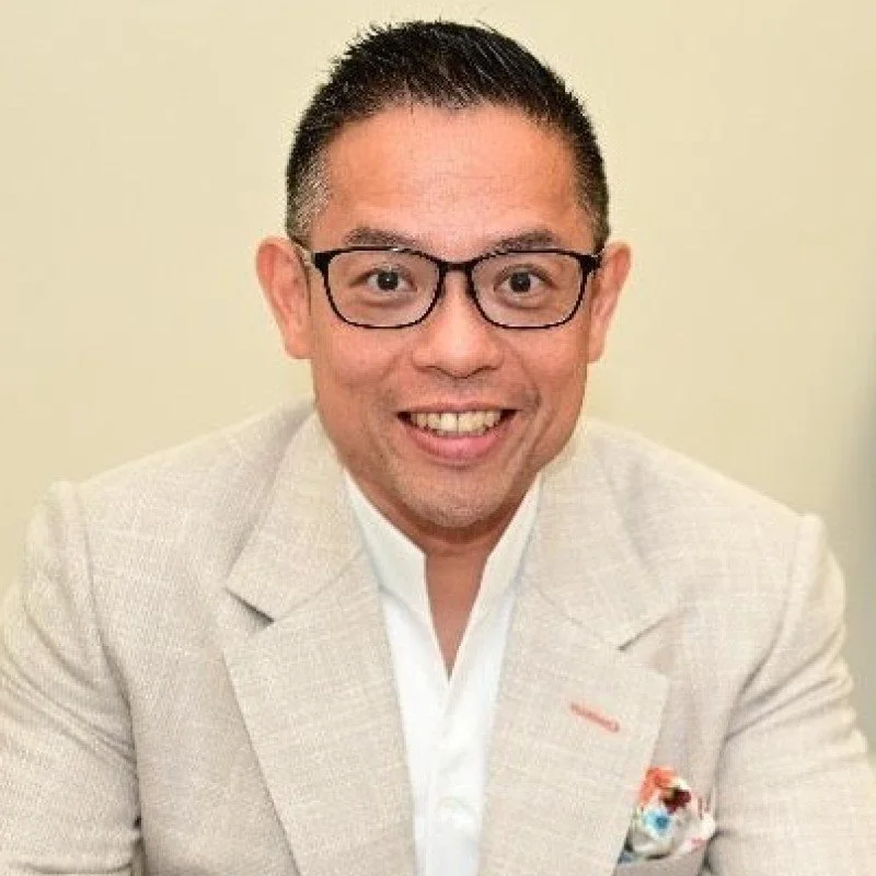 A man with glasses smiling, wearing a beige blazer and a white shirt, in front of a plain background.