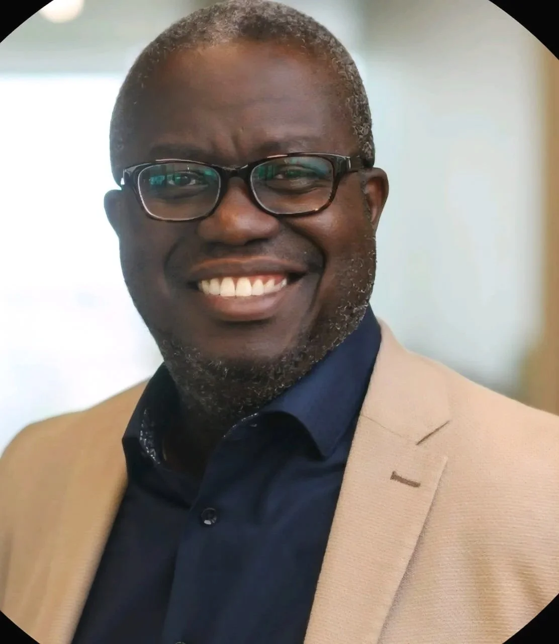 A smiling African American man with glasses, short gray hair, and a beard, wearing a beige blazer over a dark blue shirt.