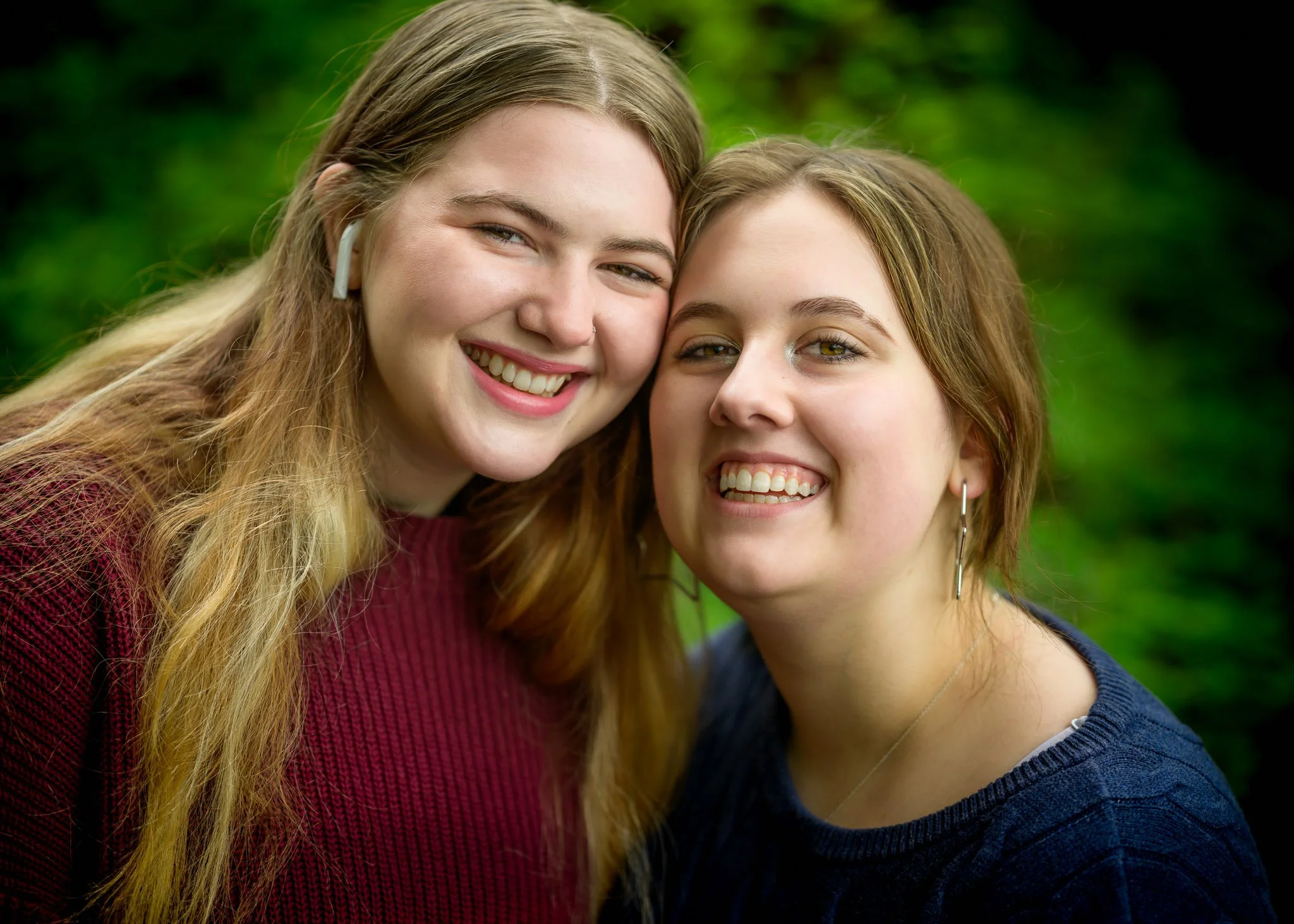 Two young women smiling closely together outdoors with green foliage in the background. Senior portraits at a park in Eugene Oregon.
