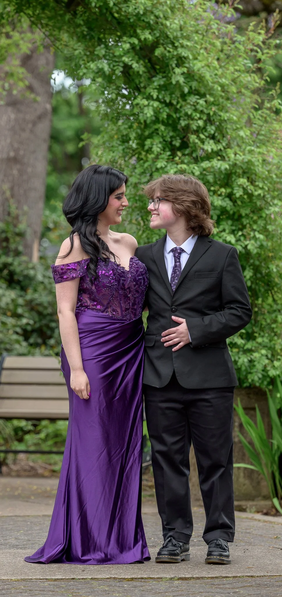 Two women in formal attire smiling and looking at each other in a garden setting, with one wearing a purple gown and the other in a black suit. Senior Prom portraits at Owen Rose Garden Eugene Oregon.
