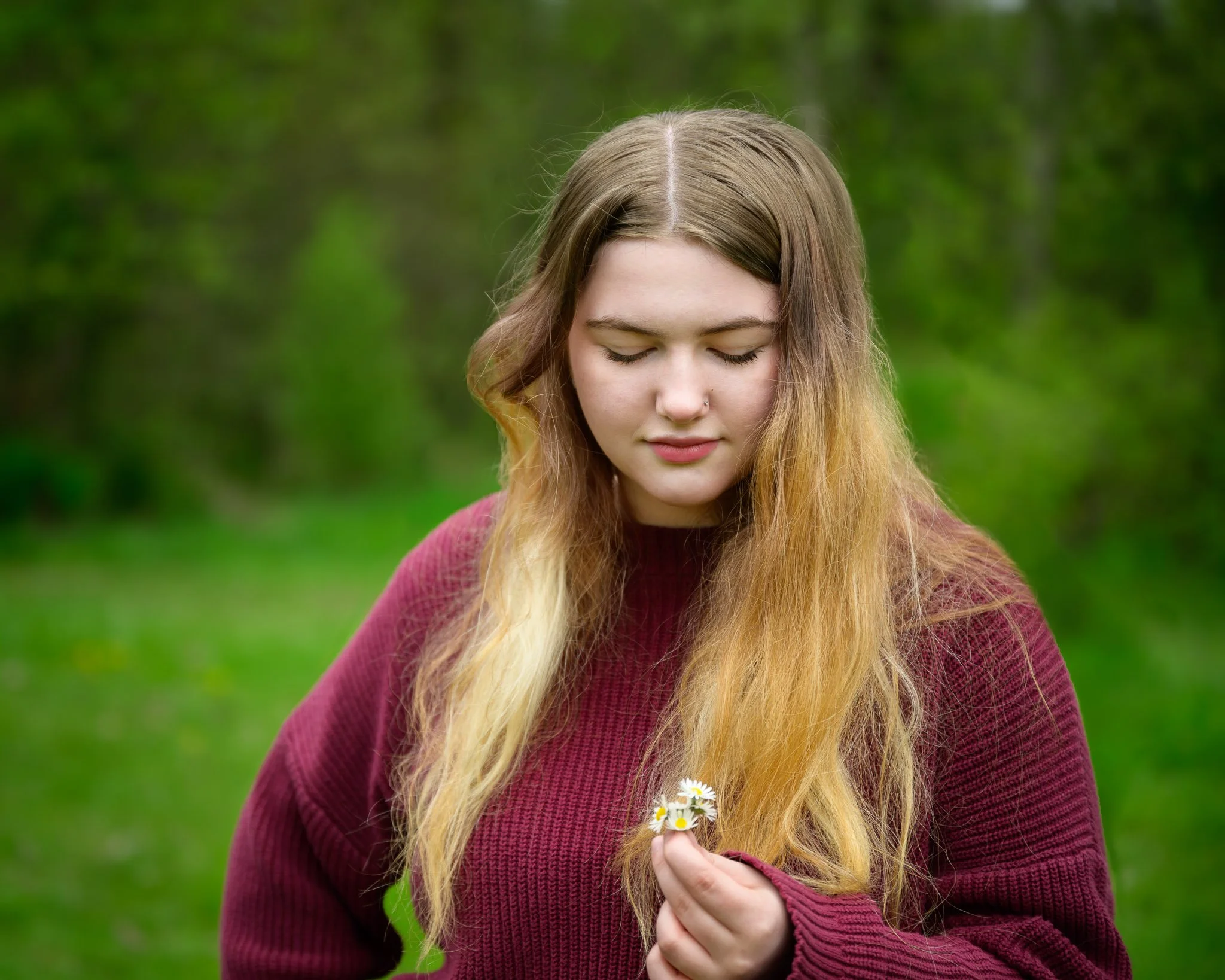 Young woman with long blonde hair holding daisies in a green outdoor setting, wearing a maroon sweater, with eyes closed. Senior portraits at a park in Eugene Oregon.