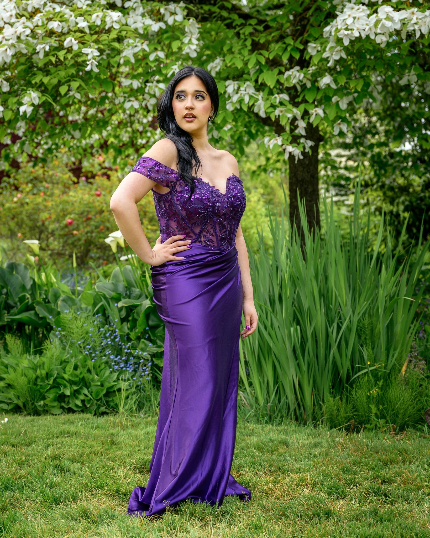 A woman wearing a purple evening gown standing outdoors in a garden with green plants and white flowering trees in the background. Senior Prom portraits at Owen Rose Garden Eugene Oregon.