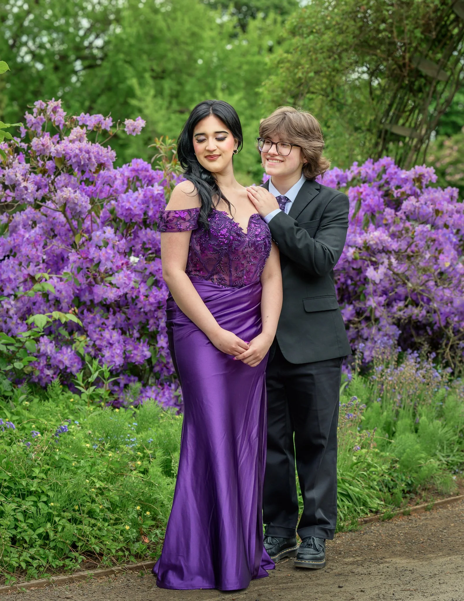 A young woman in an elegant purple gown and a young man in a black suit with glasses pose together in front of blooming purple flowers outdoors. Senior Prom portraits at Owen Rose Garden Eugene Oregon.