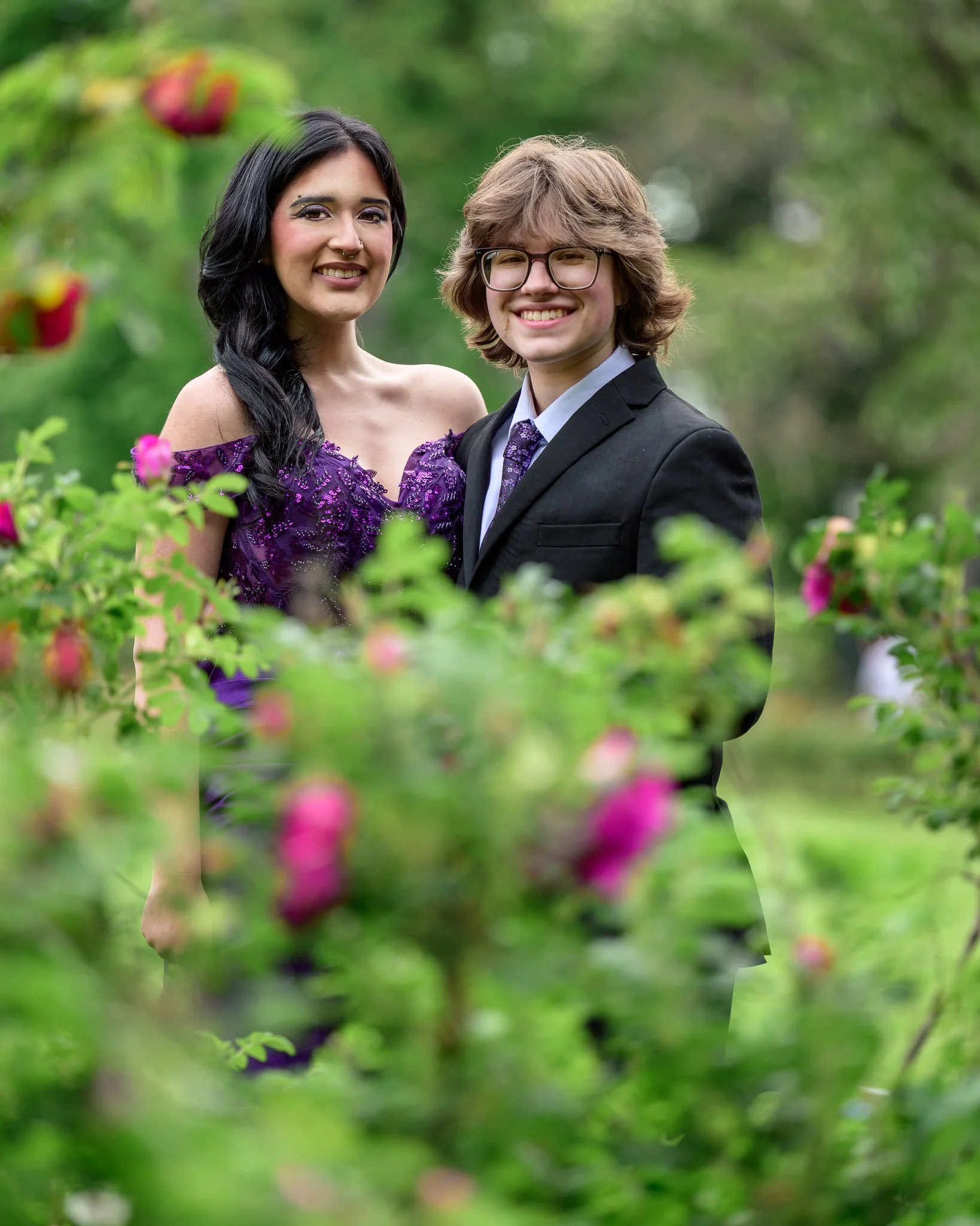 A young woman in a purple dress and a young man in a black suit with glasses are standing outdoors among green foliage and pink flowers, smiling at the camera. Senior Prom portraits at Owen Rose Garden Eugene Oregon.