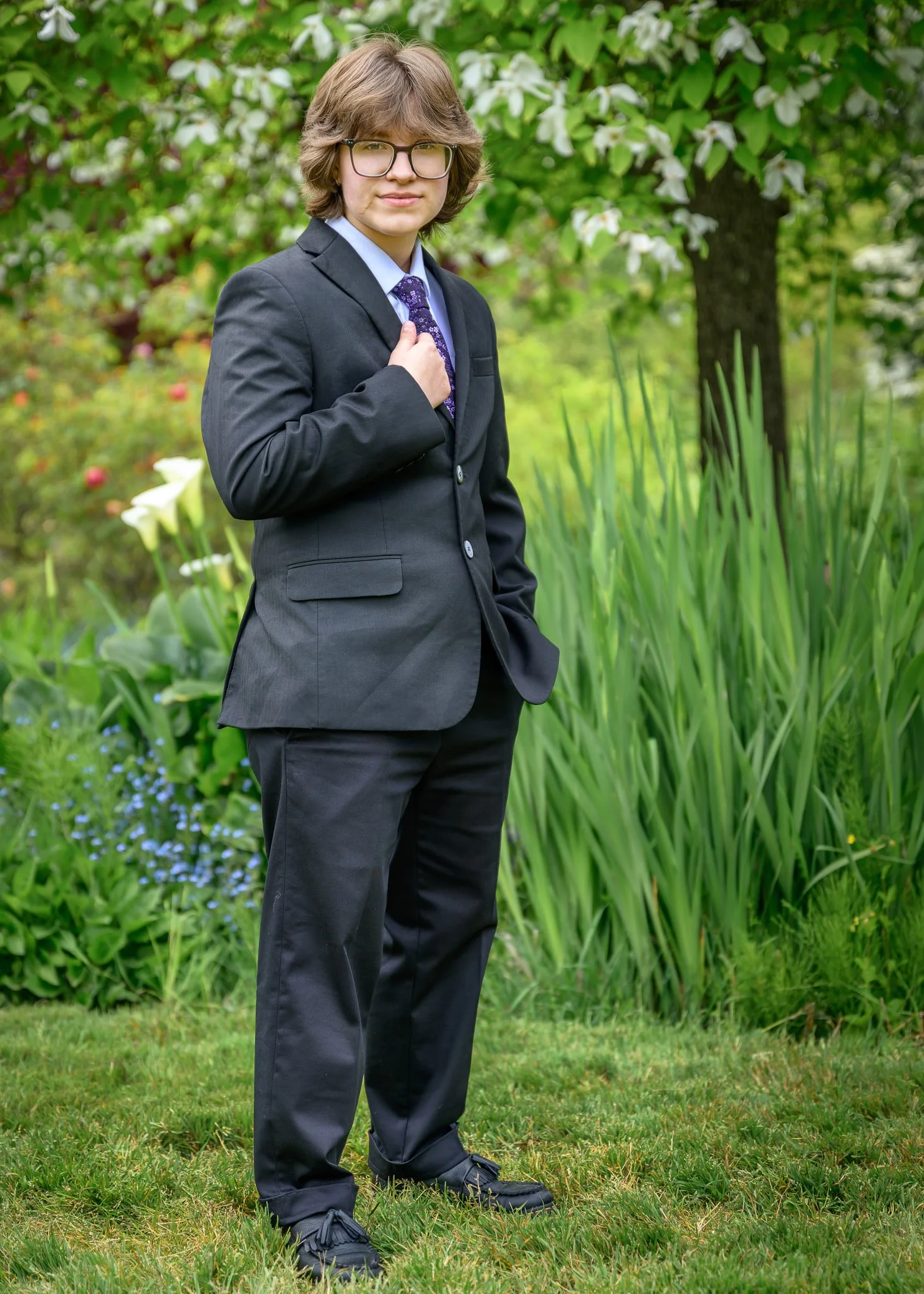 A young person with brown hair and glasses wearing a black suit and tie standing outdoors in a garden with green plants and trees. Senior Prom portraits at Owen Rose Garden Eugene Oregon.