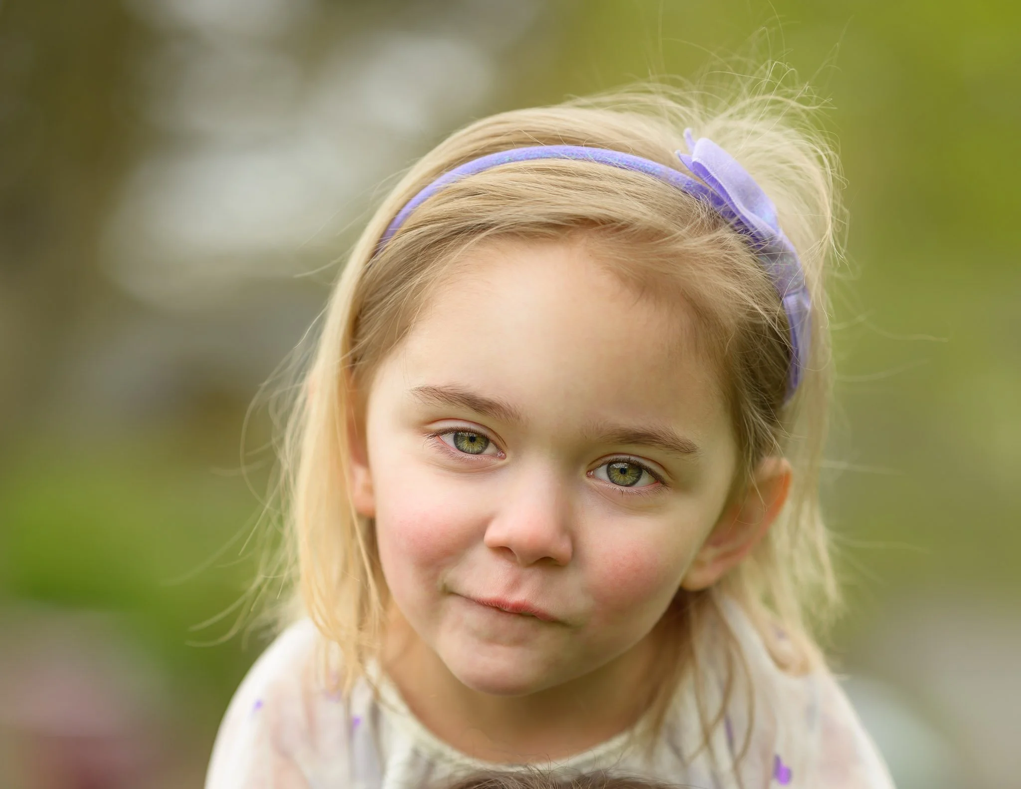 Close-up of a young girl with blonde hair, green eyes, and a purple headband outdoors with blurred greenery in the background. Natural light family photography in Eugene Oregon.