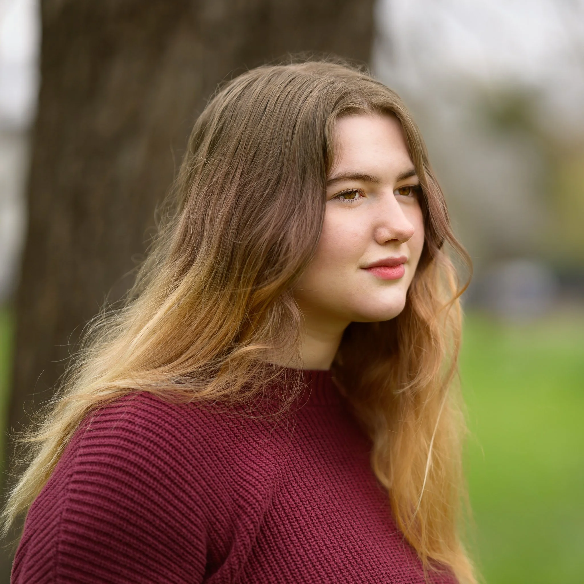 A young woman with long, wavy brown hair standing outdoors near a tree, wearing a maroon sweater. Senior portraits at a park in Eugene Oregon.