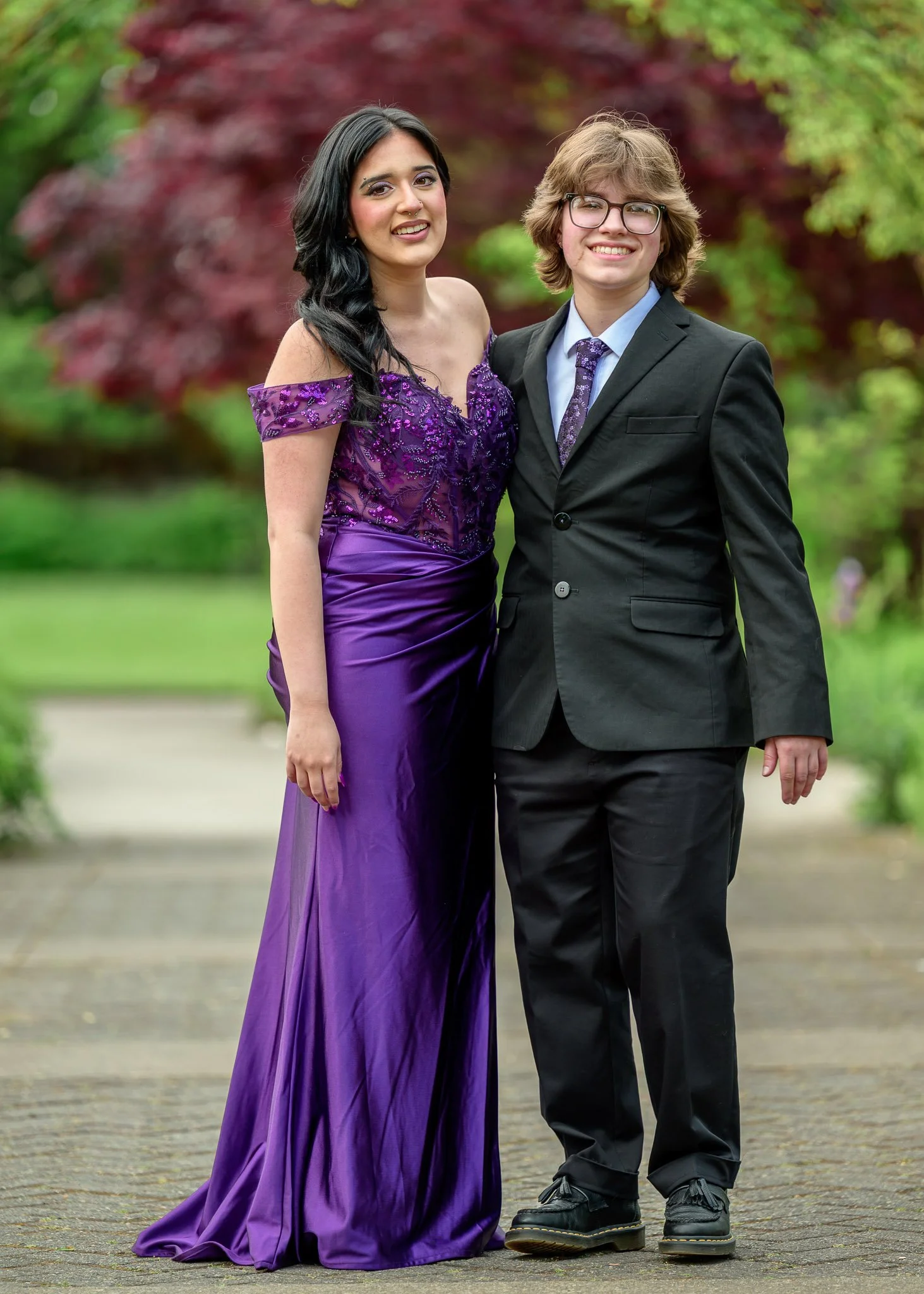 A young woman in a purple gown and a young man in a black suit with a purple tie standing together outdoors in a park with greenery and a red-leafed tree in the background. Senior Prom portraits at Owen Rose Garden Eugene Oregon.