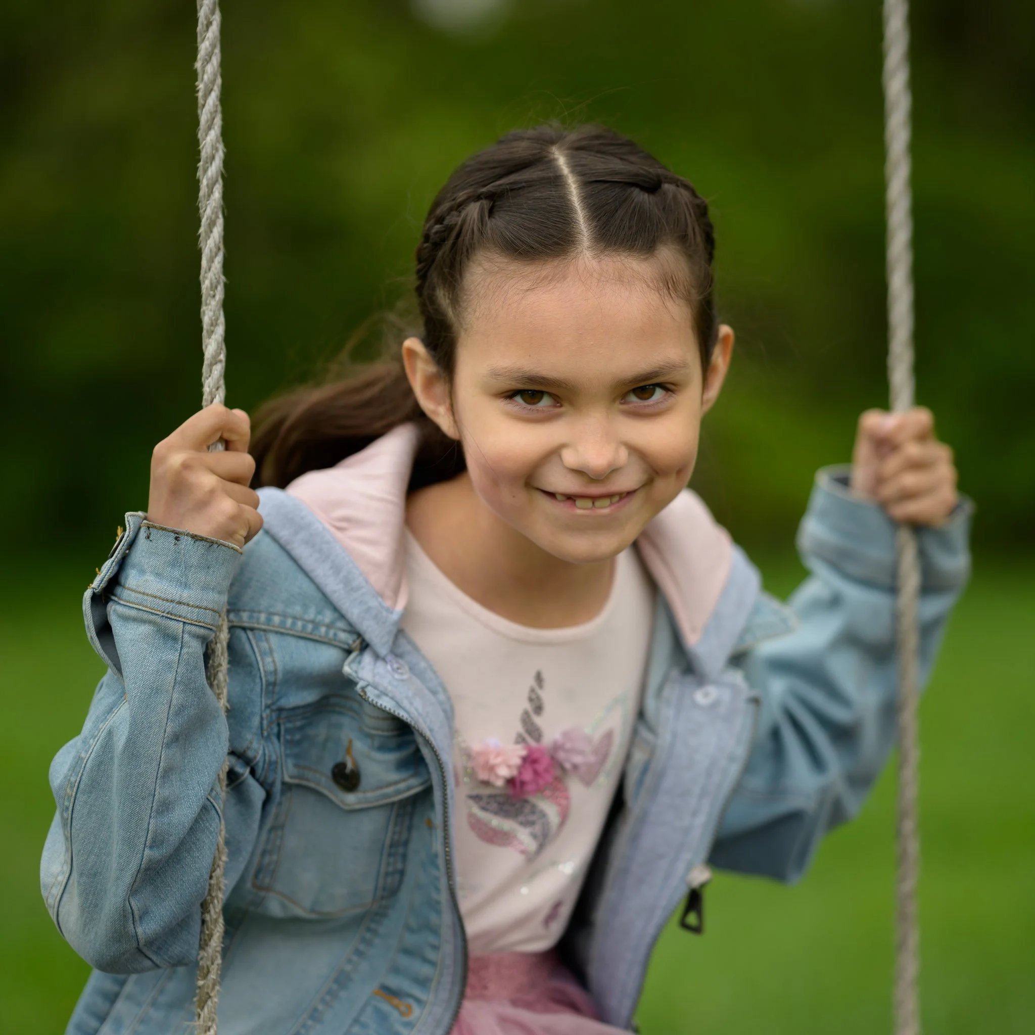 Young girl with brown hair in braids smiling and holding onto playground swing ropes outdoors. Family photography at local park Eugene Oregon.
