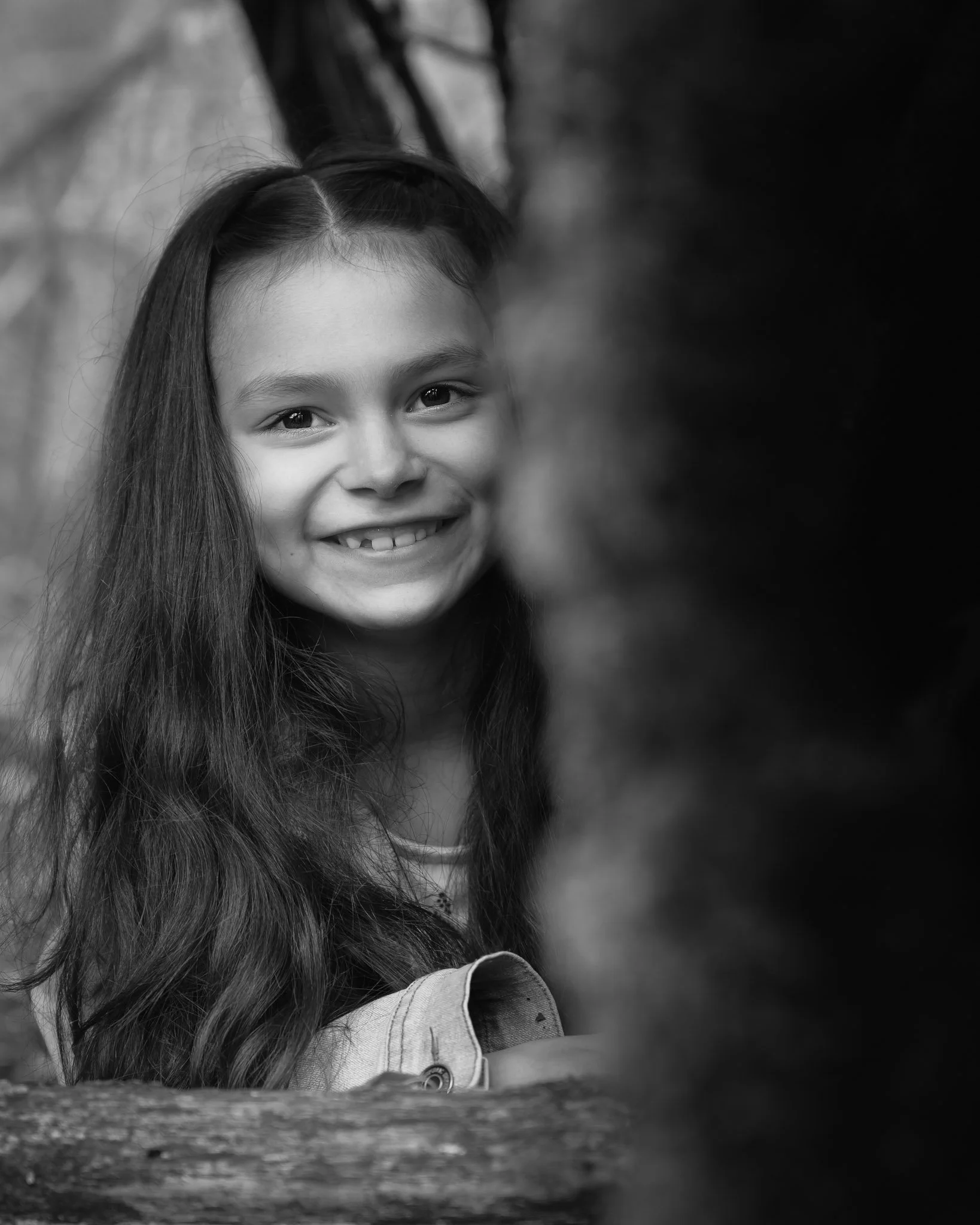 A smiling young girl with long hair looking directly at the camera in an outdoor setting, black and white photo. Family photography at local park Eugene Oregon.