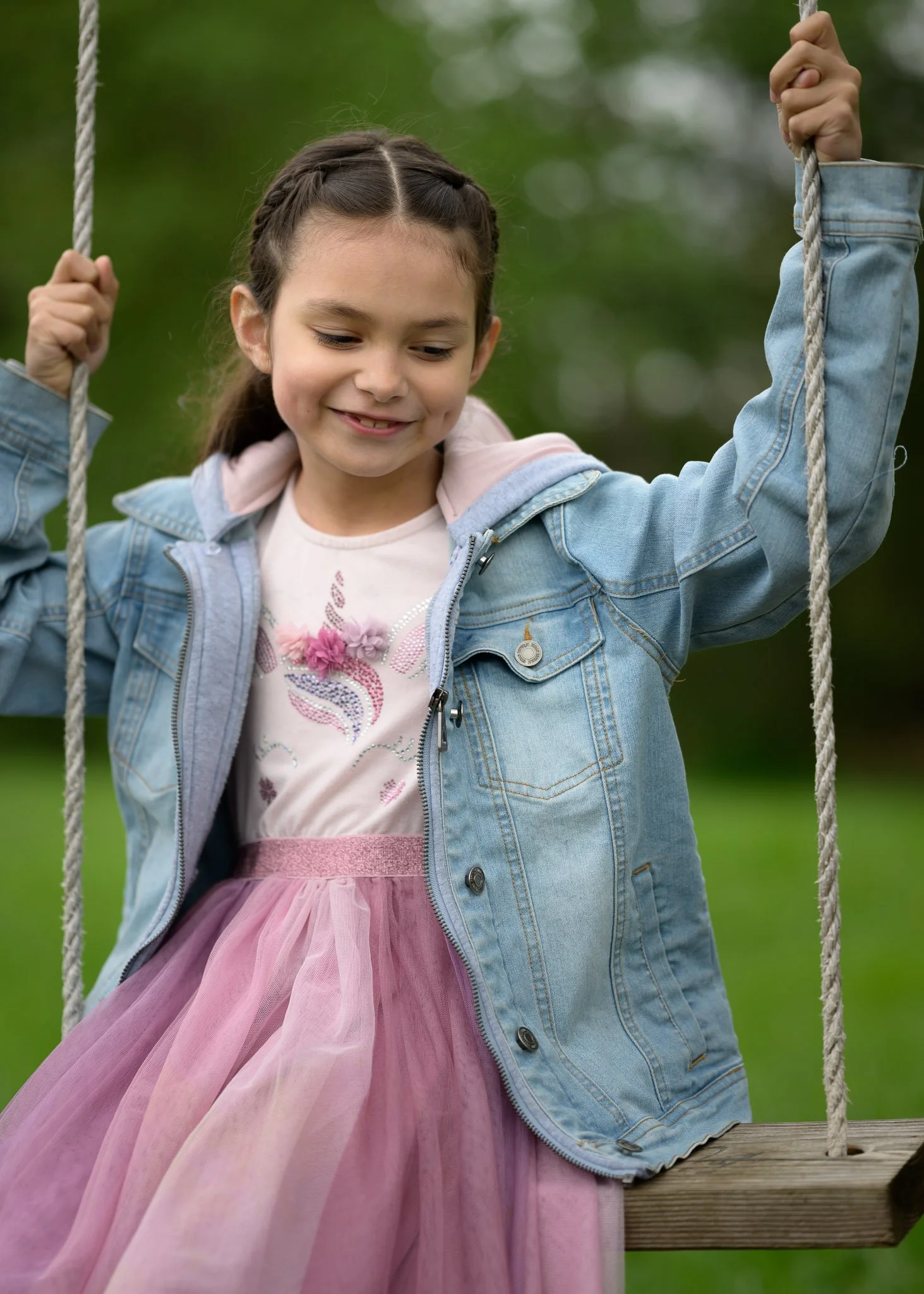 Young girl in a denim jacket and pink tutu skirt smiling on a wooden swing outdoors. Family photography at local park Eugene Oregon.