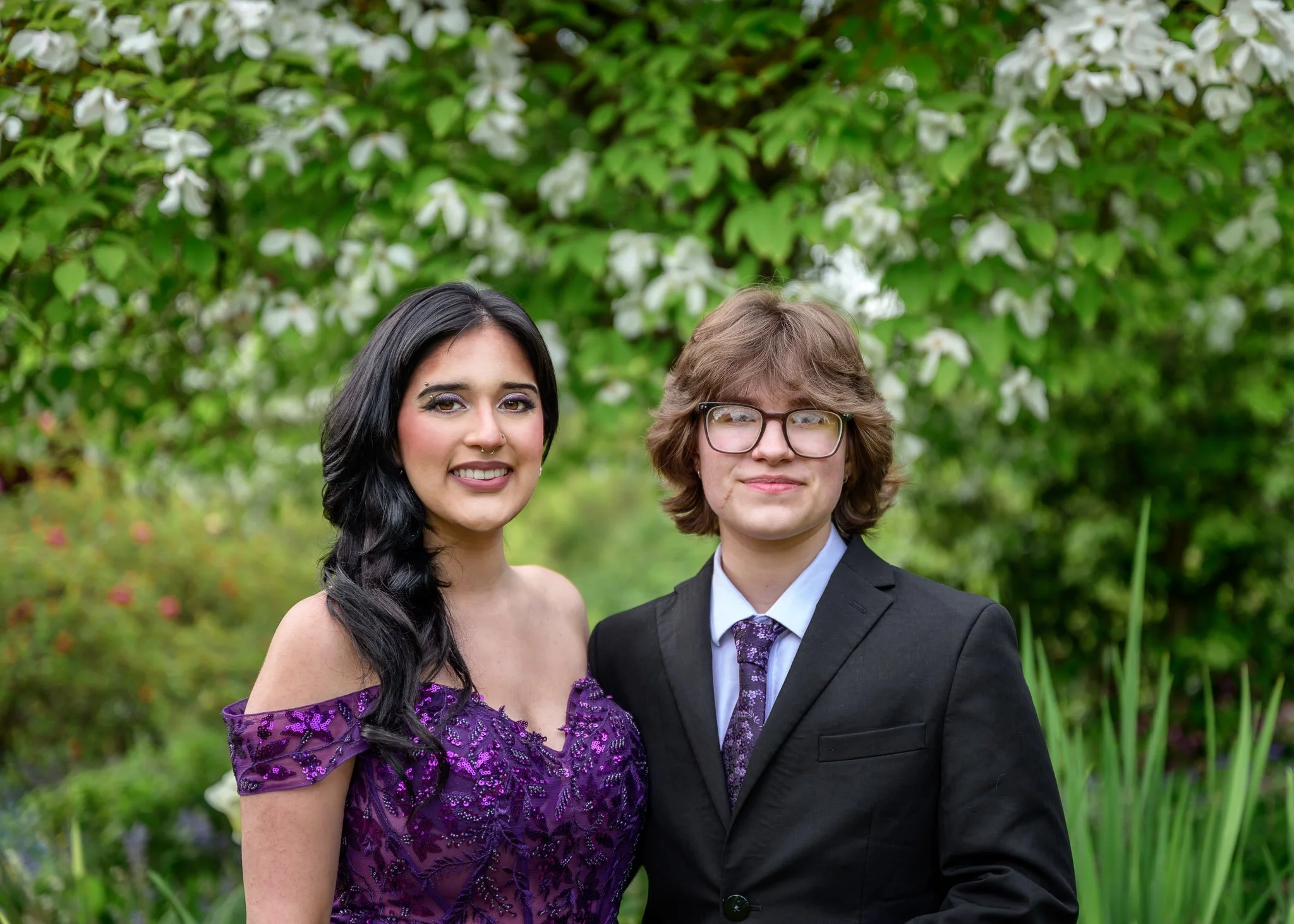 A young woman in a purple dress and a young man in a black suit with glasses standing outdoors in front of green bushes with white flowers. Senior Prom portraits at Owen Rose Garden Eugene Oregon.