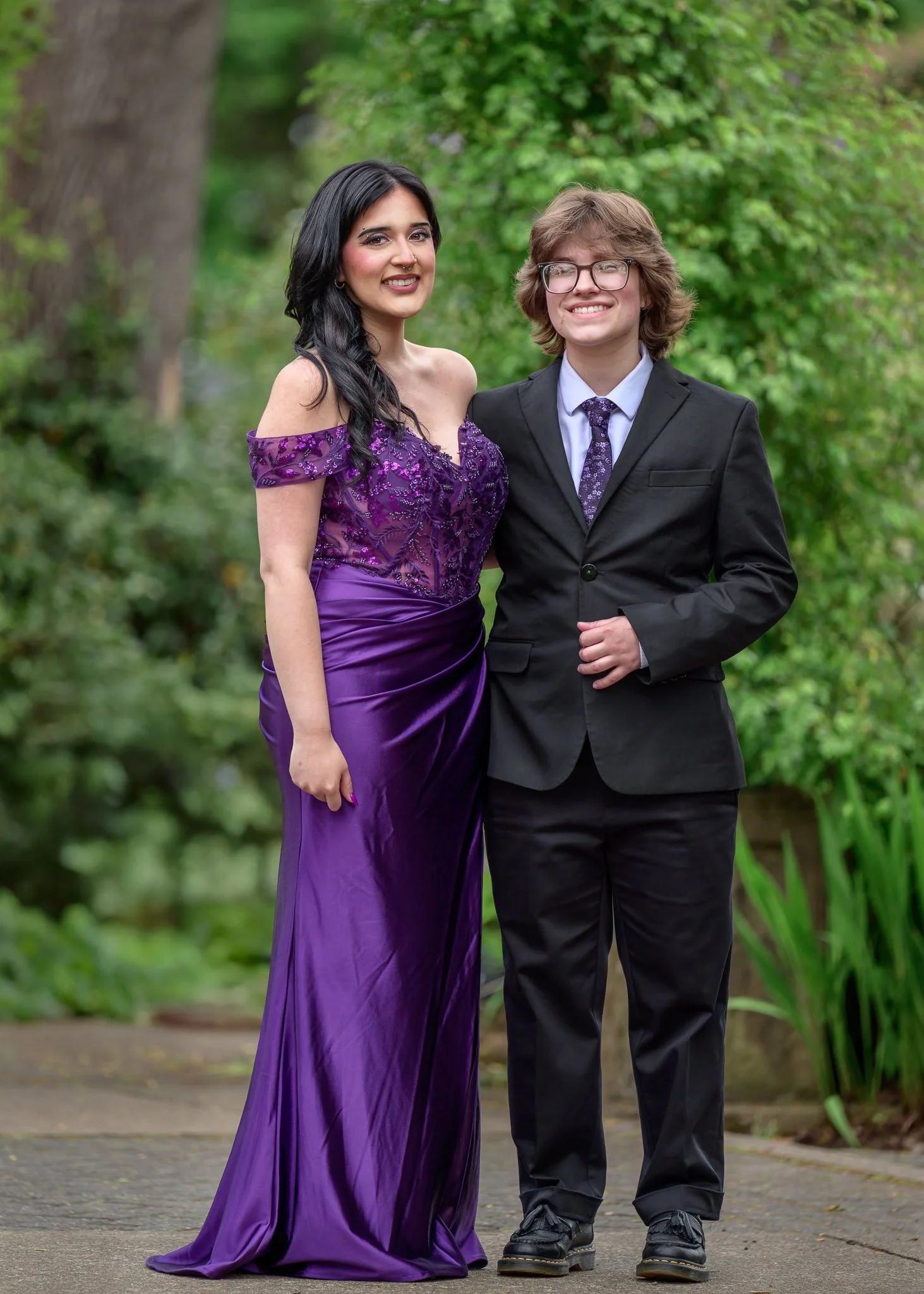 A woman in a purple gown and a young man in a black suit with glasses standing together outdoors with green trees in the background. Senior Prom portraits at Owen Rose Garden Eugene Oregon.