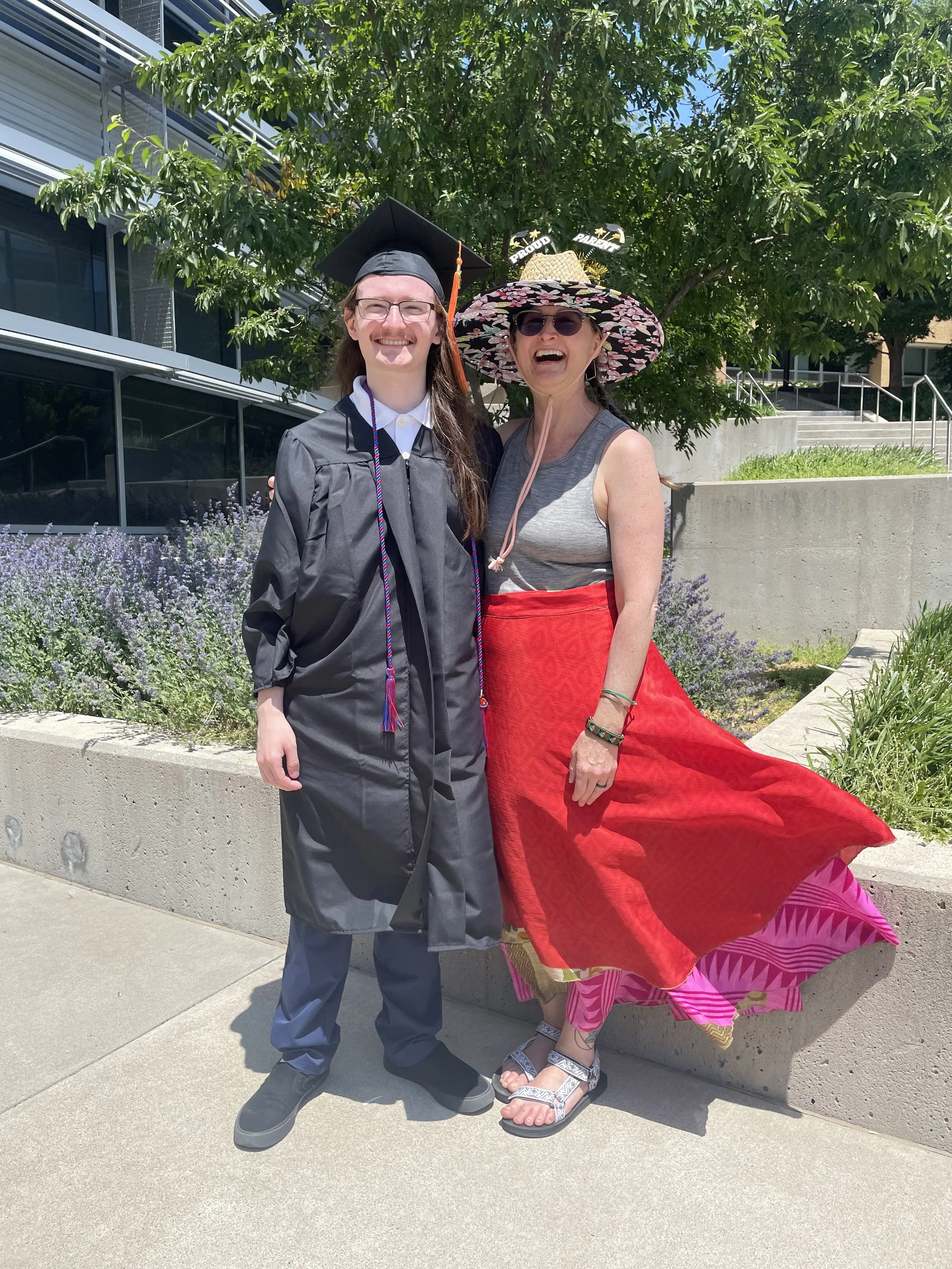 A young man in a black graduation gown and cap standing next to an older woman in a colorful hat and red skirt, outdoors on a sunny day.