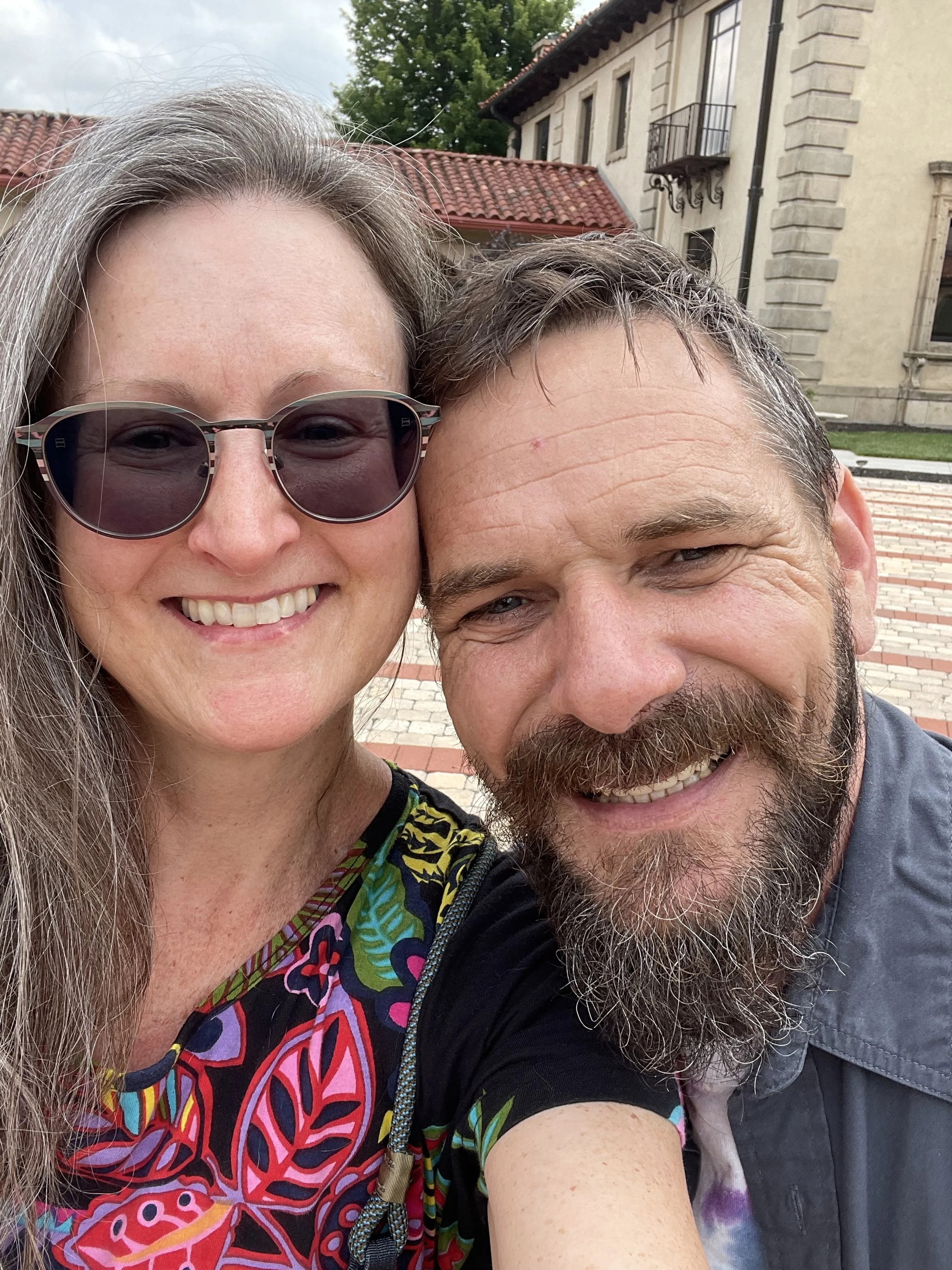 Close-up of a smiling woman with long gray hair wearing sunglasses and a colorful patterned top, and a smiling man with a beard and short hair, outdoors in front of a building with a red-tiled roof and a paved walkway.