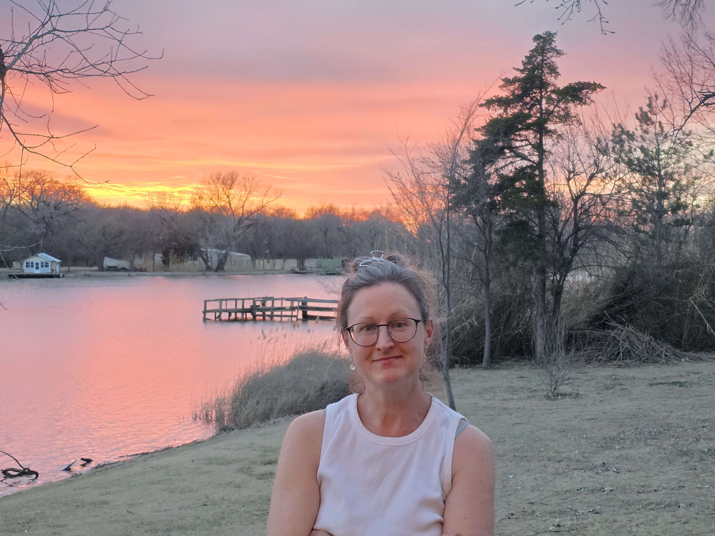 A woman with glasses and a white sleeveless top standing outdoors near a lake during sunset, with trees and a small dock in the background.