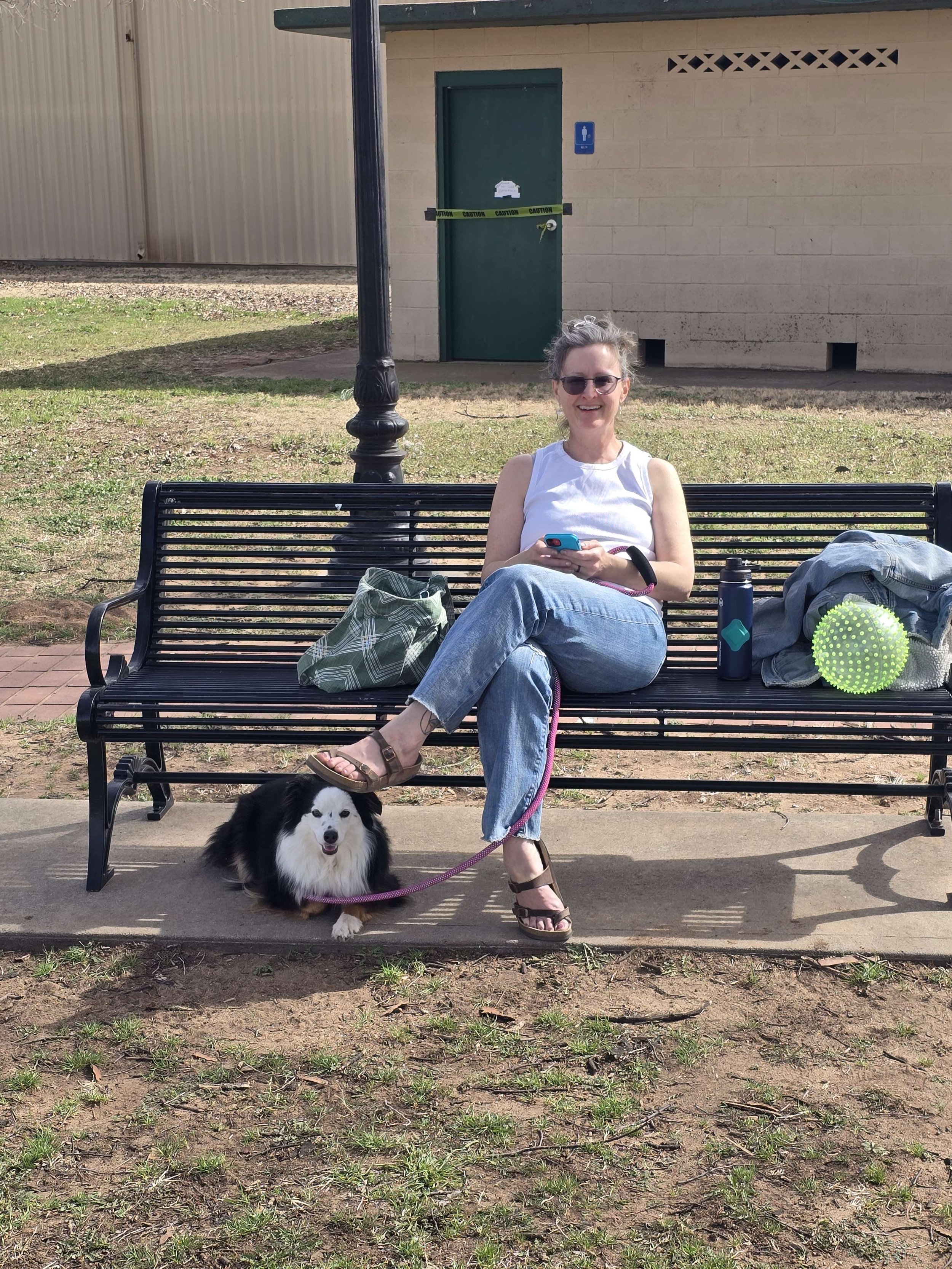 A woman with gray hair, sunglasses, white sleeveless top, and jeans sitting on a park bench, holding a phone, with a small black-and-white dog lying on the ground beside her leash. The bench has personal items, including a water bottle, backpack, and a green textured ball, and is situated on a concrete pad with grass and a building with a green door in the background.
