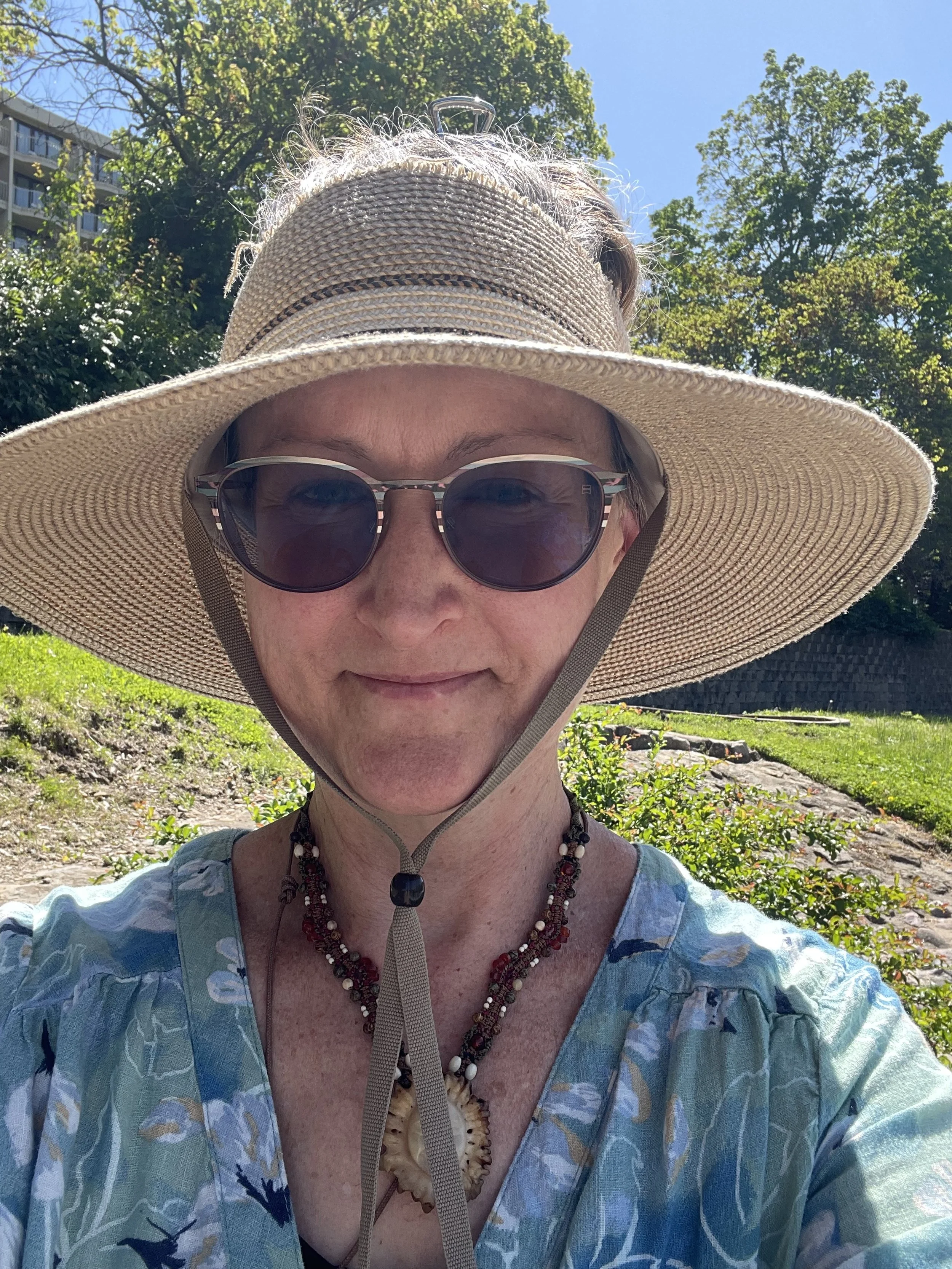 A woman wearing a wide-brimmed straw hat, sunglasses, a floral blouse, and beaded necklaces in a sunny outdoor setting with trees and greenery in the background.