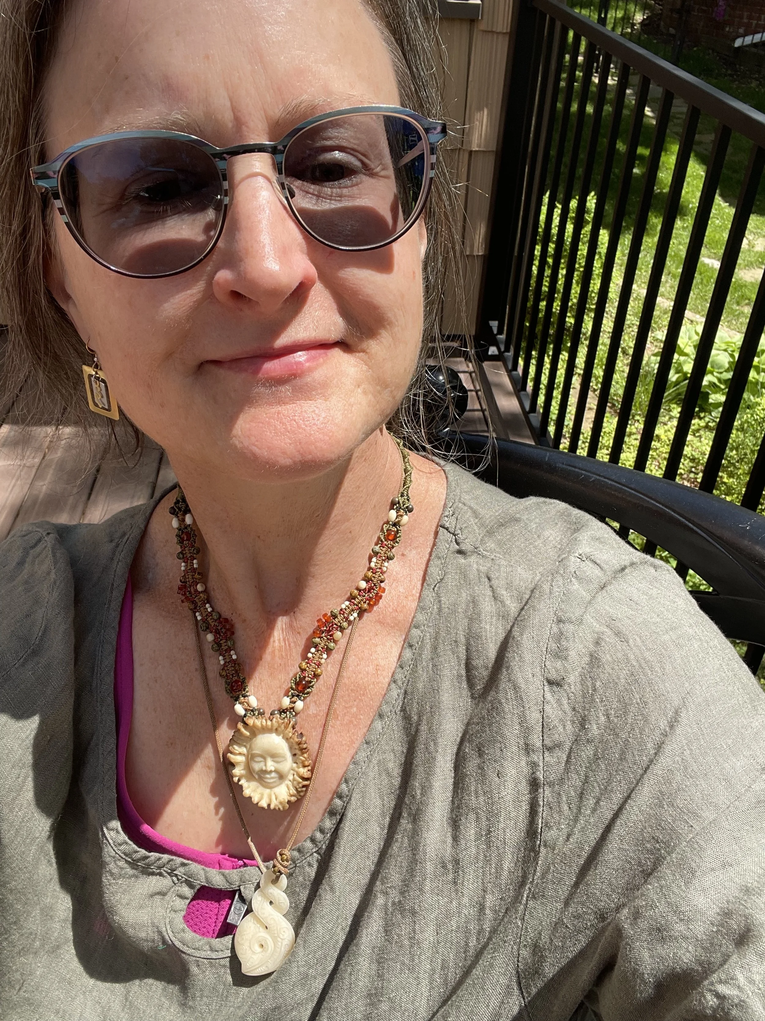 A woman wearing sunglasses, earrings, and multiple necklaces, sitting outdoors on a sunny day, with a wooden deck and green foliage in the background.
