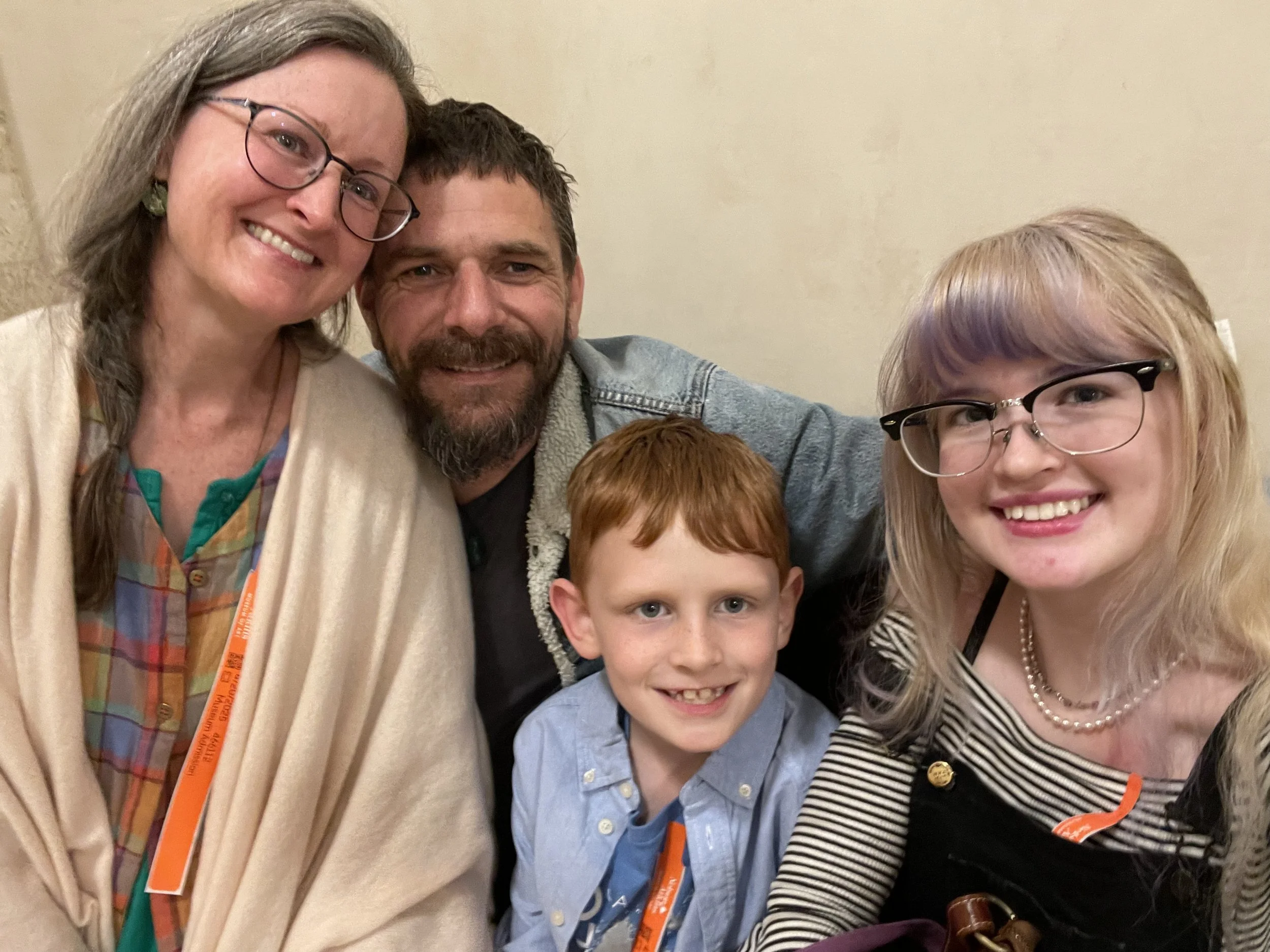 A smiling multi-generational family of four taking a selfie indoors, including a woman with glasses, a man with a beard, a young boy, and a young woman with glasses.