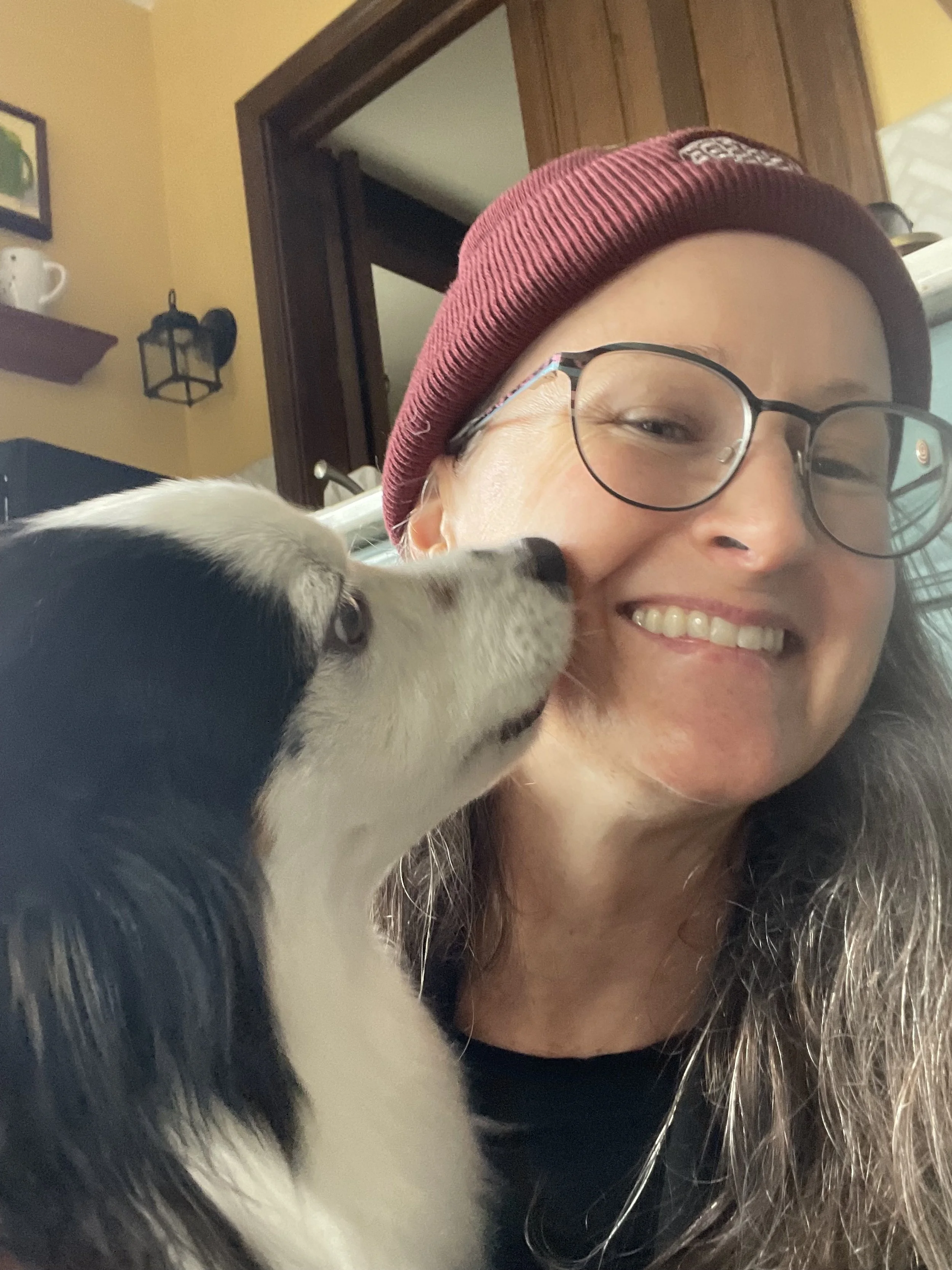 A woman smiling and wearing glasses and a red beanie, with a black and white puppy licking her face.
