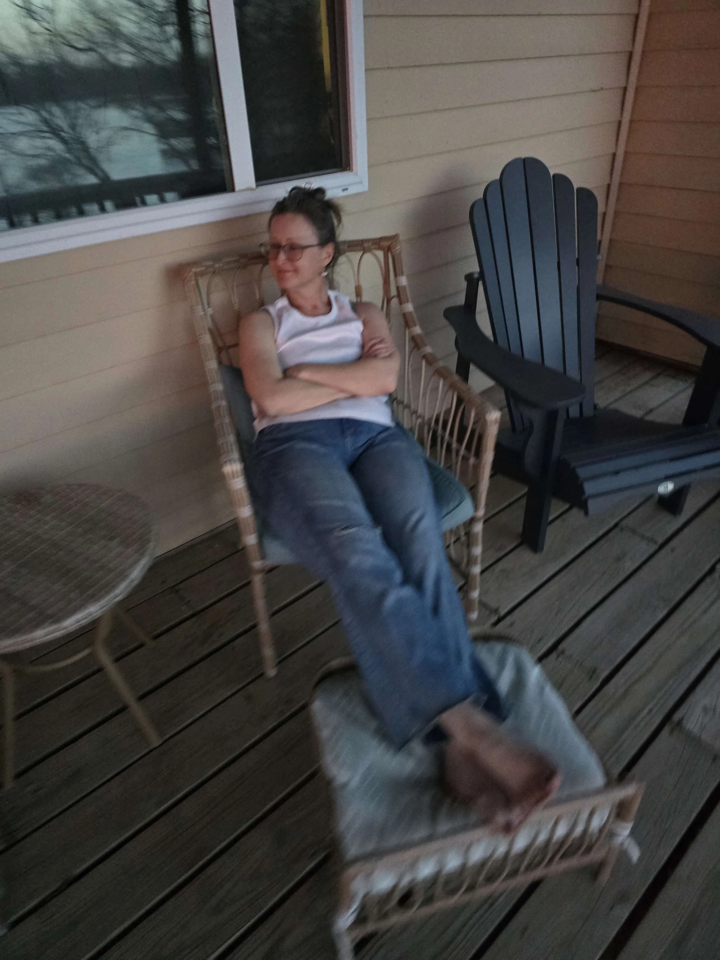 A woman sitting on a wicker chair on a wooden porch, with her arms crossed and smiling, next to a black Adirondack chair and a small round table.