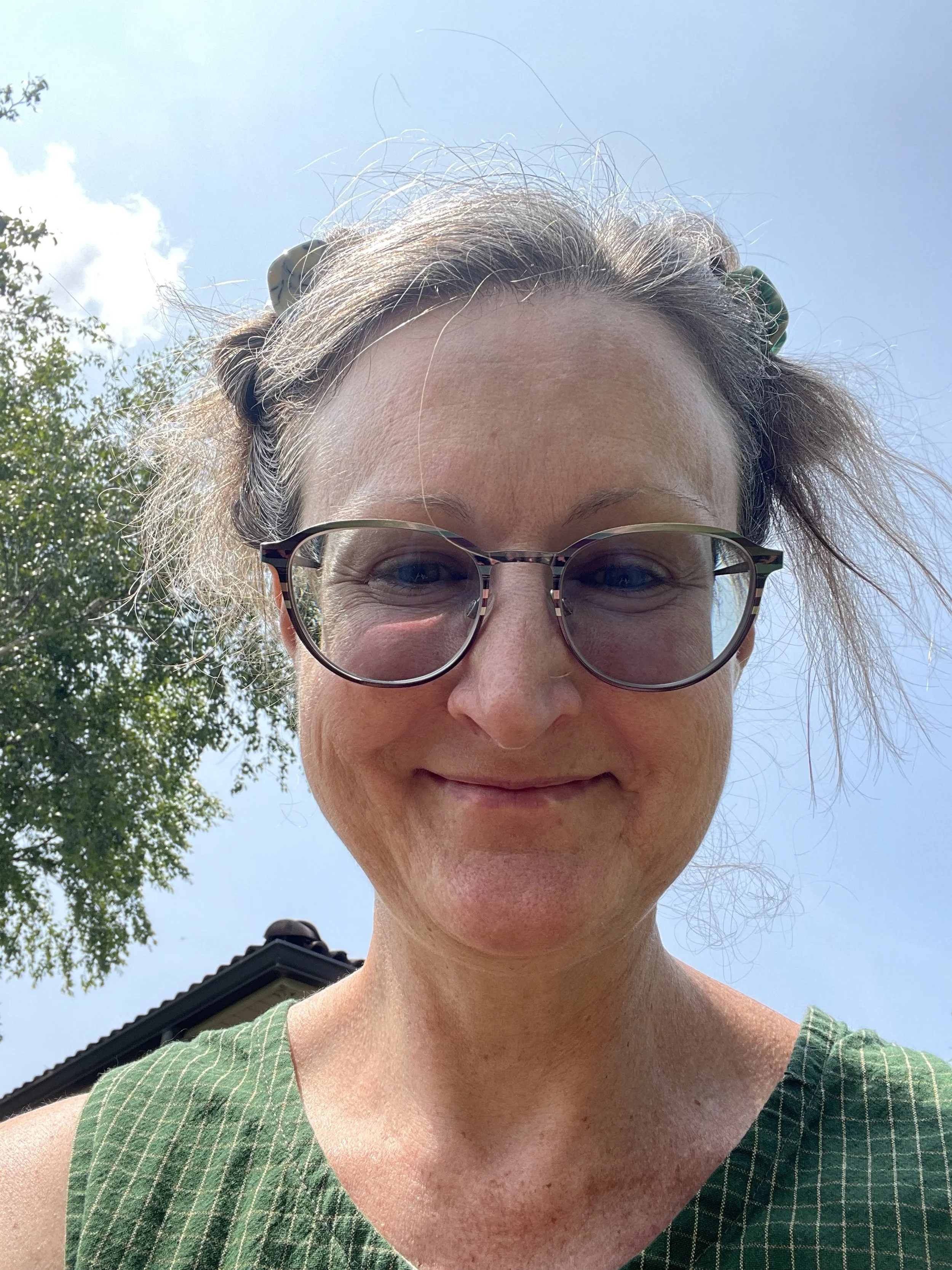 Close-up of a woman wearing glasses, smiling outdoors on a sunny day, with a clear blue sky and a tree visible in the background.