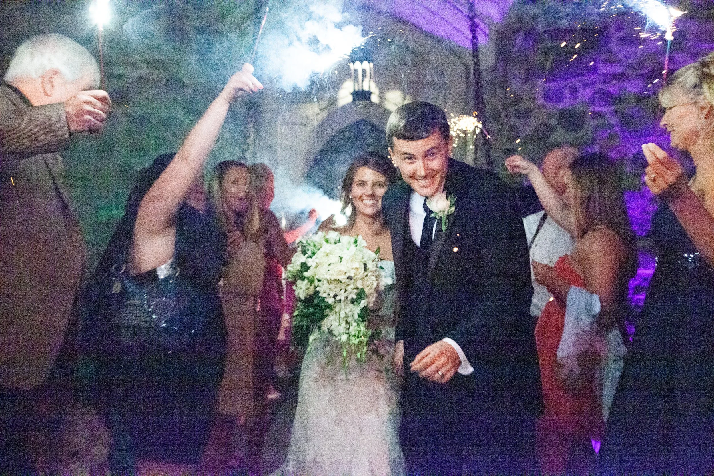 A joyful wedding celebration with a bride and groom surrounded by guests, sparklers, and colorful lighting.