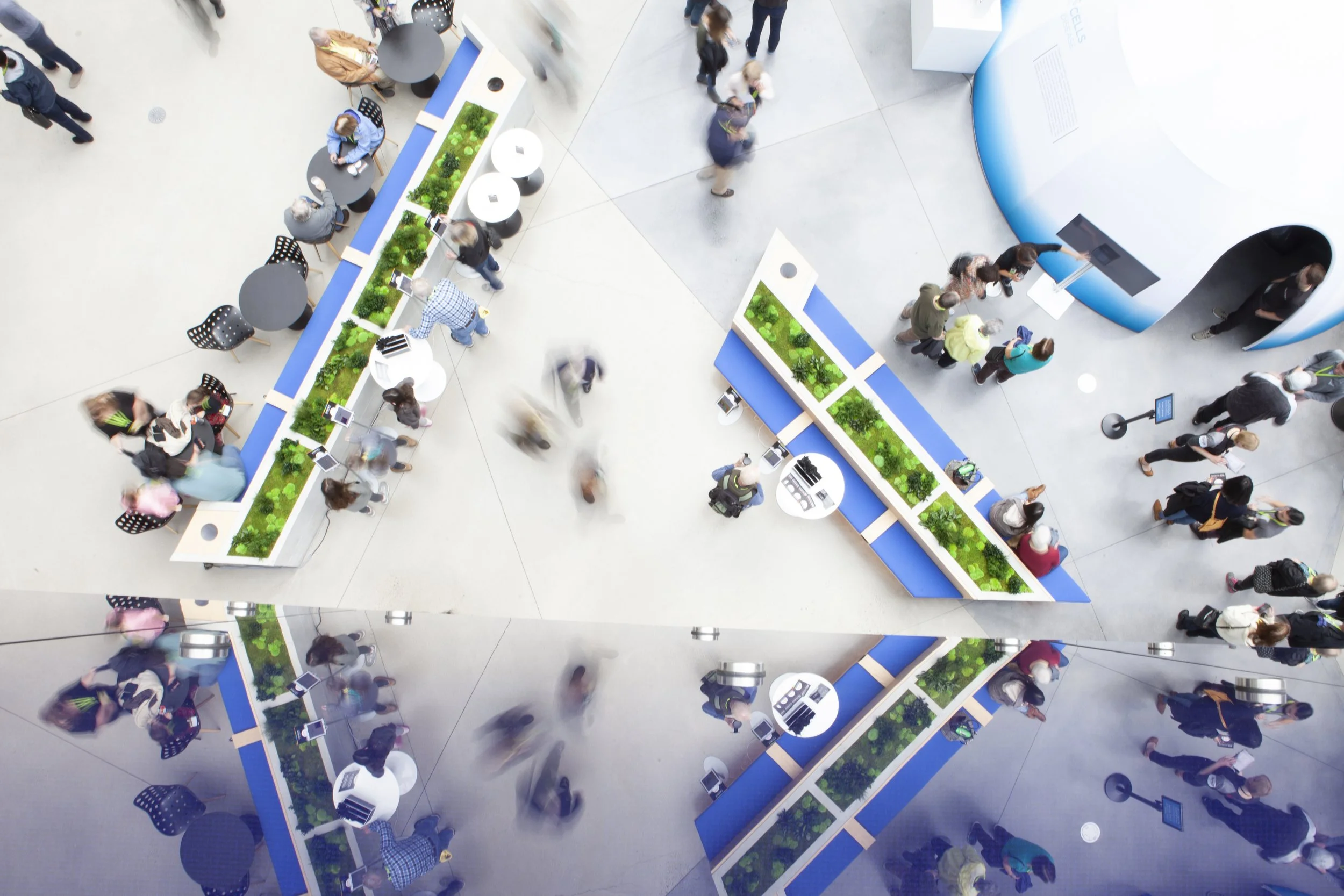 Aerial view of a modern indoor setting with people walking and standing around tables and planters. The space features blue counters with green plants and reflective surfaces beneath. Small groups are interacting, creating a bustling atmosphere.