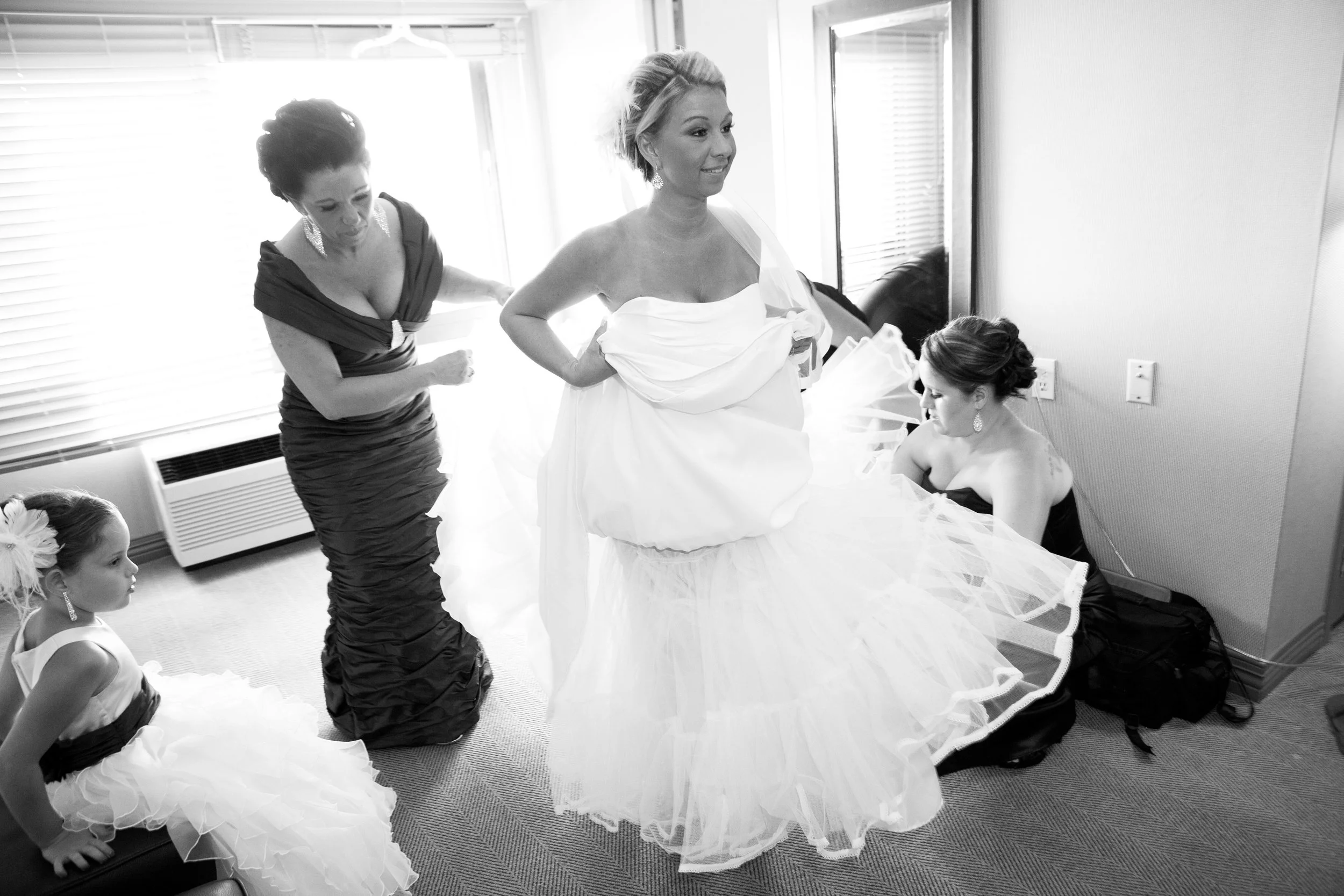 Women and a girl preparing for a wedding; one woman is in a wedding dress, others assisting her.