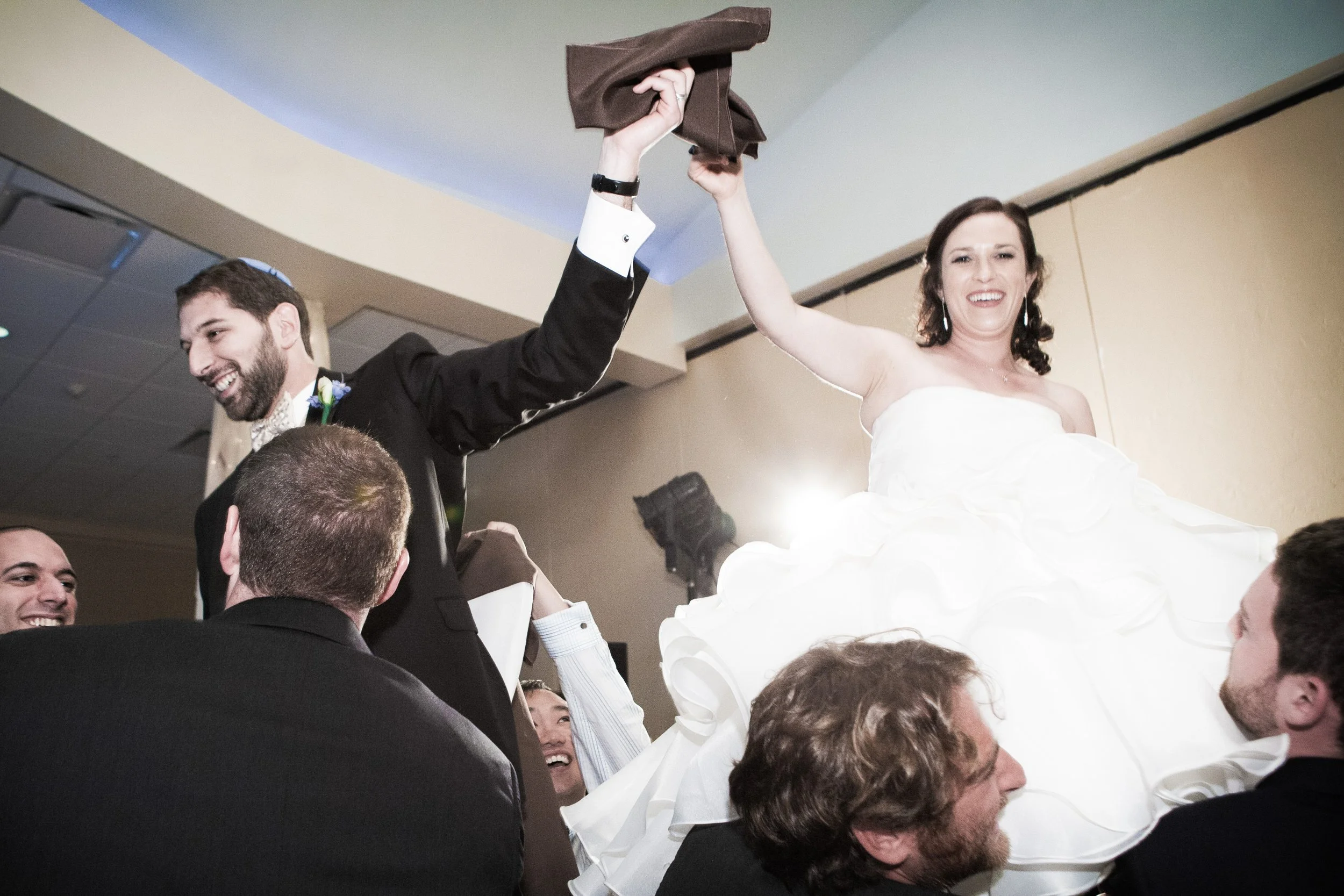 Bride and groom being lifted at a jewish wedding celebration, smiling and holding hands.