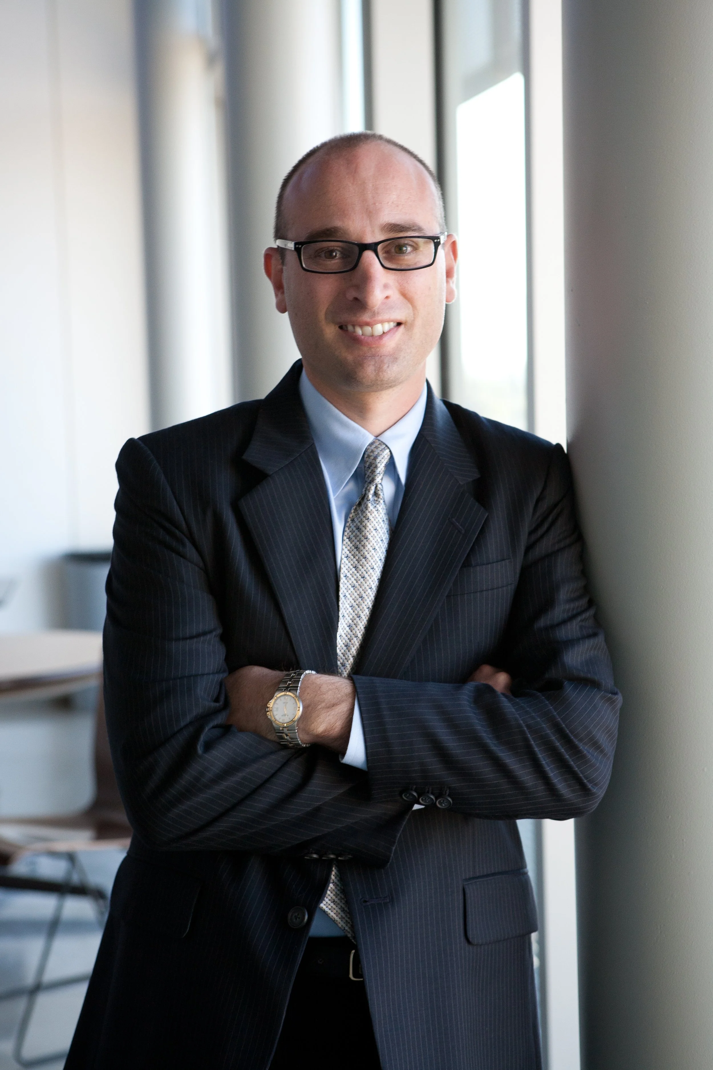 Man in a suit and glasses leaning against a wall, smiling
