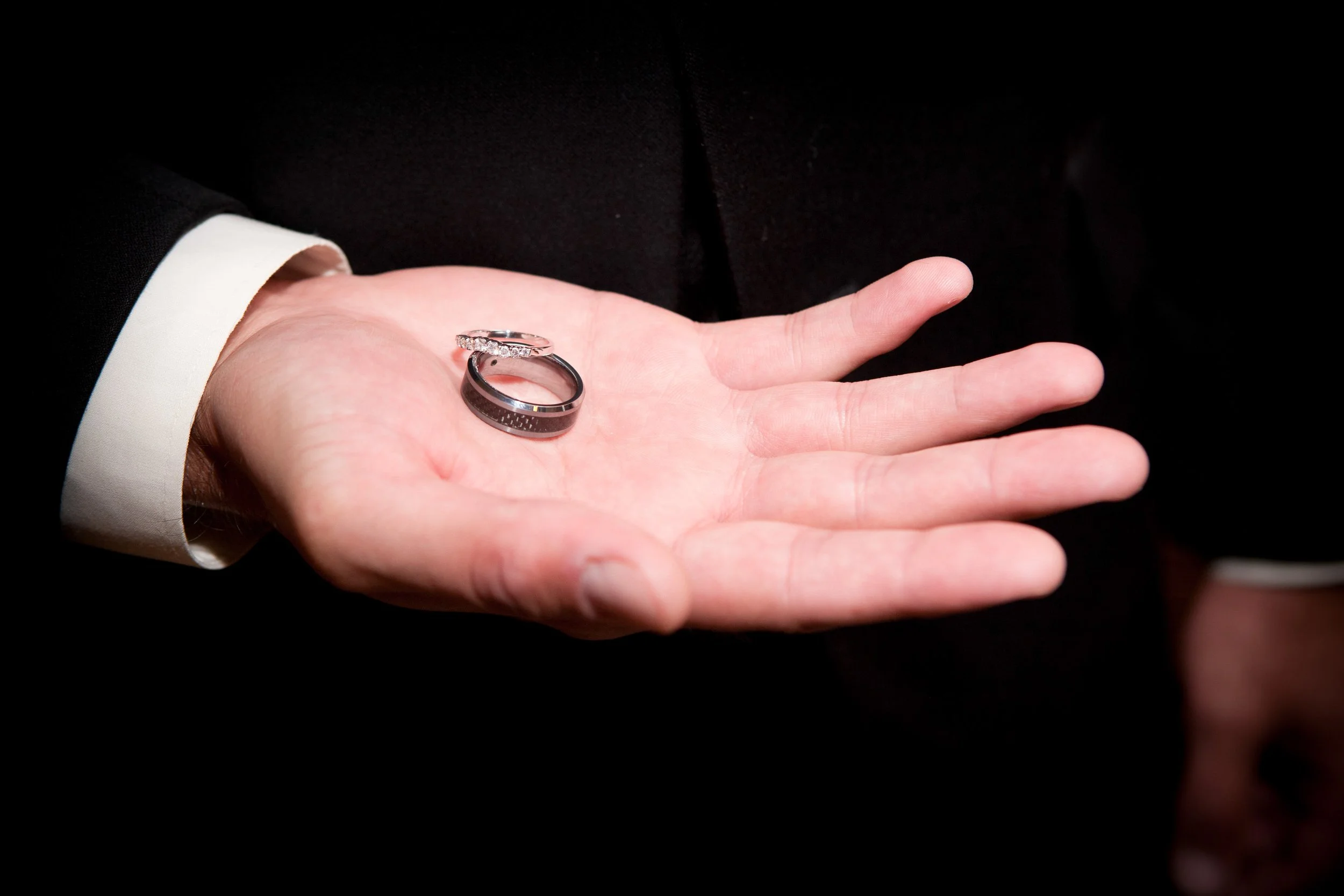 A groom in a black suit holding two wedding rings in his hand. One ring is silver with embedded diamonds, and the other is a black band.
