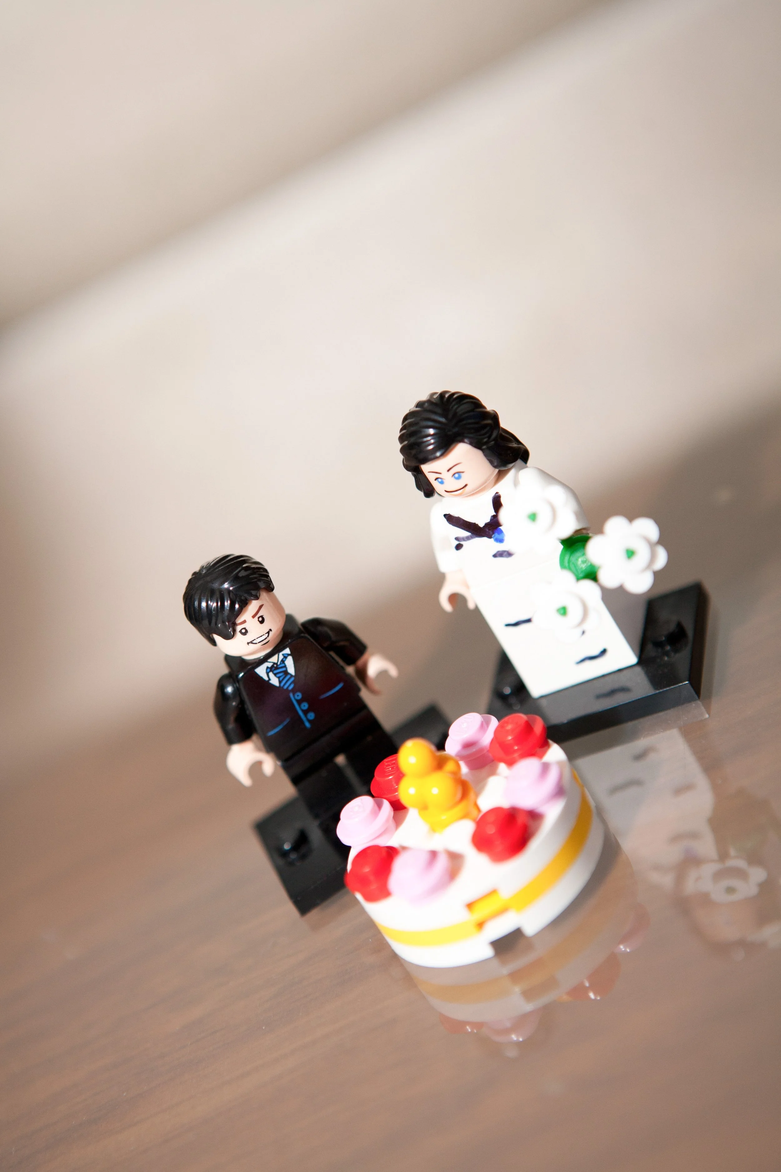 Lego figurines of a bride and groom in wedding attire standing on a table with a cake decorated with cherries and pink flowers.