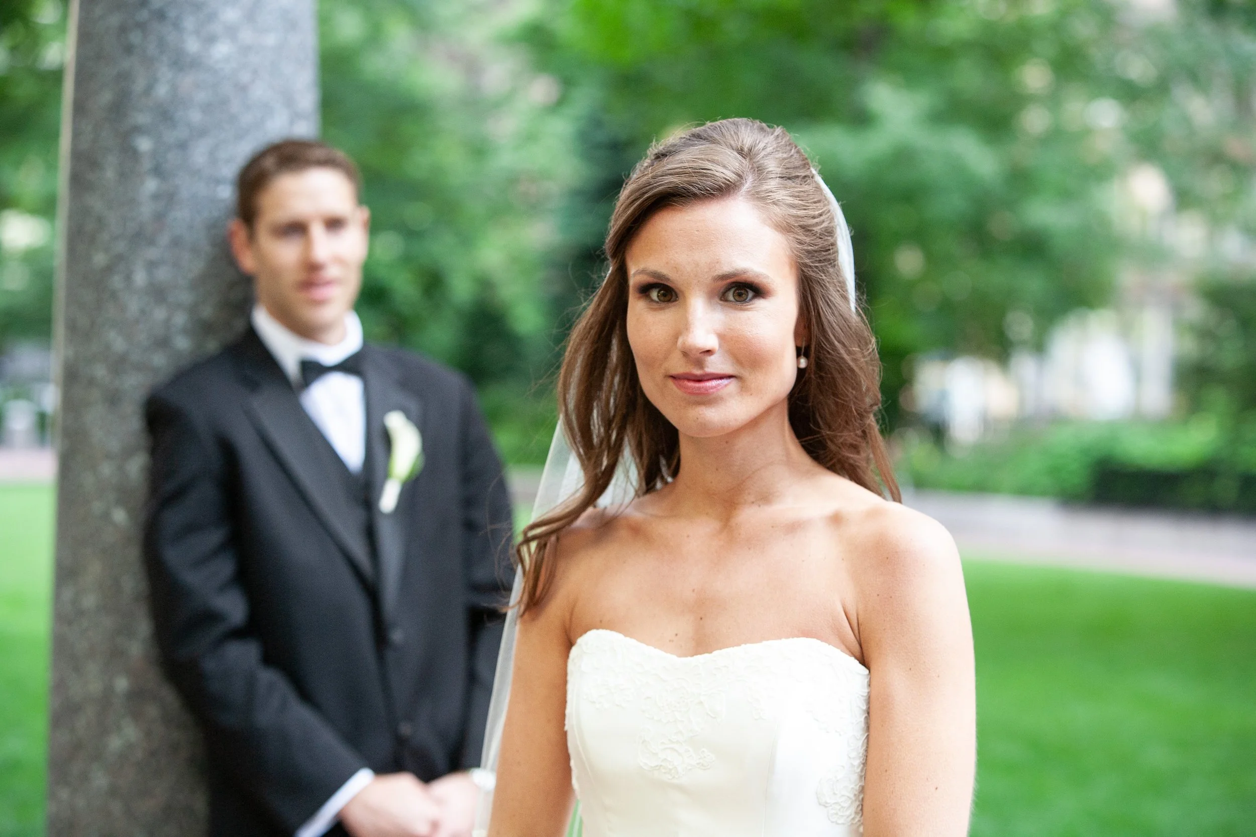 A bride in a white wedding dress standing outdoors with a groom in a black tuxedo blurred in the background, near a tree and green park setting.