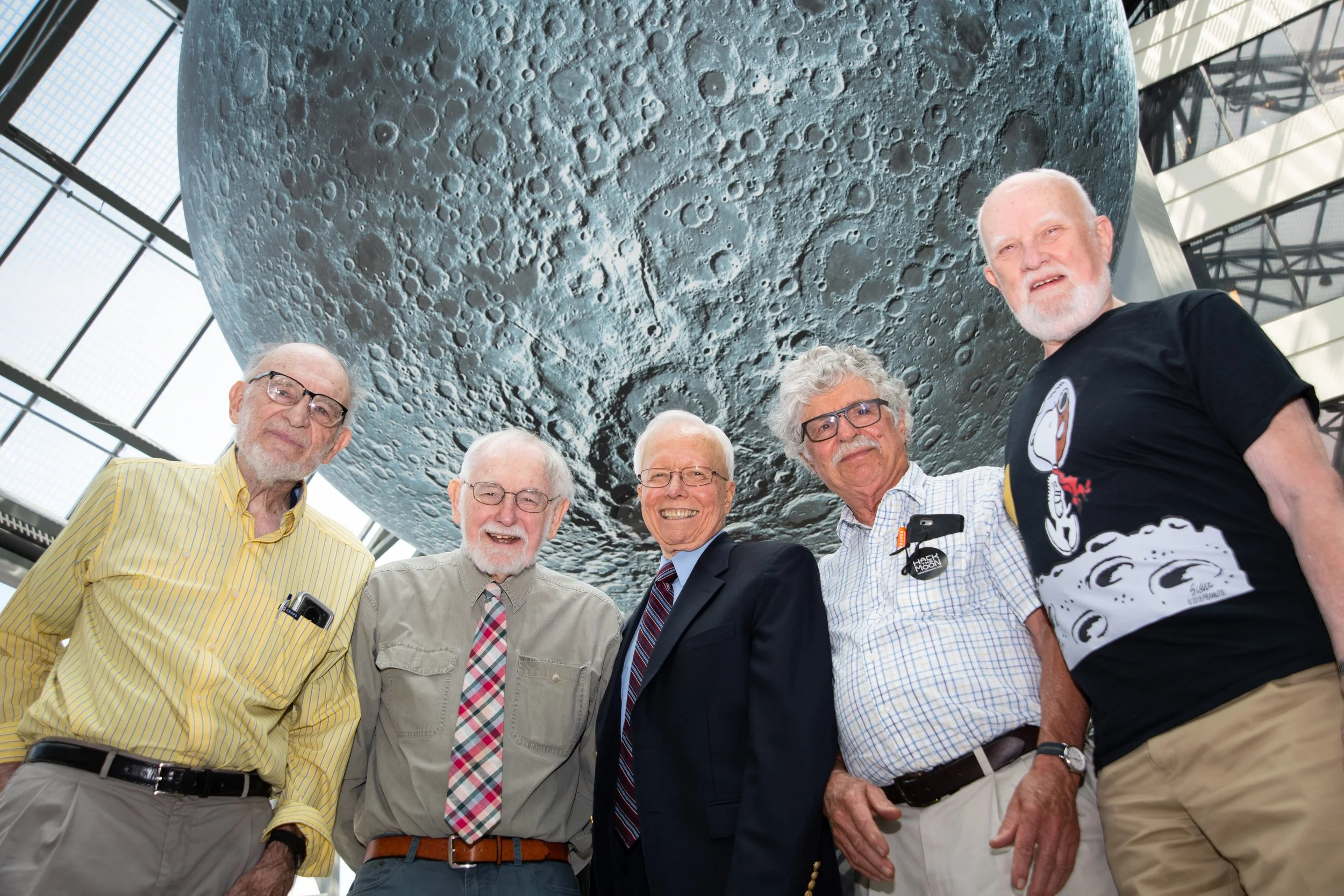 Five elderly men standing under a large moon model inside a building with a glass ceiling.