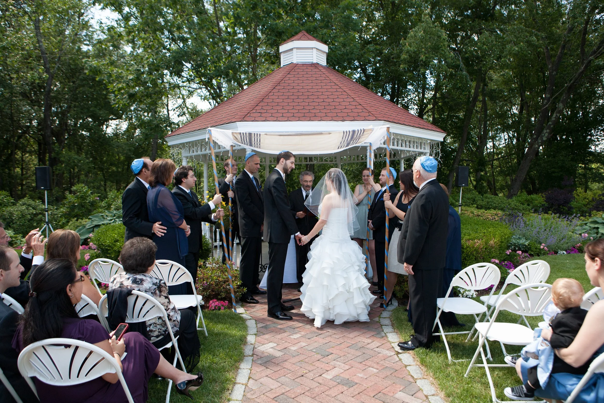 Wedding ceremony with bride and groom holding hands in an outdoor garden, surrounded by guests, under a gazebo, during daytime.