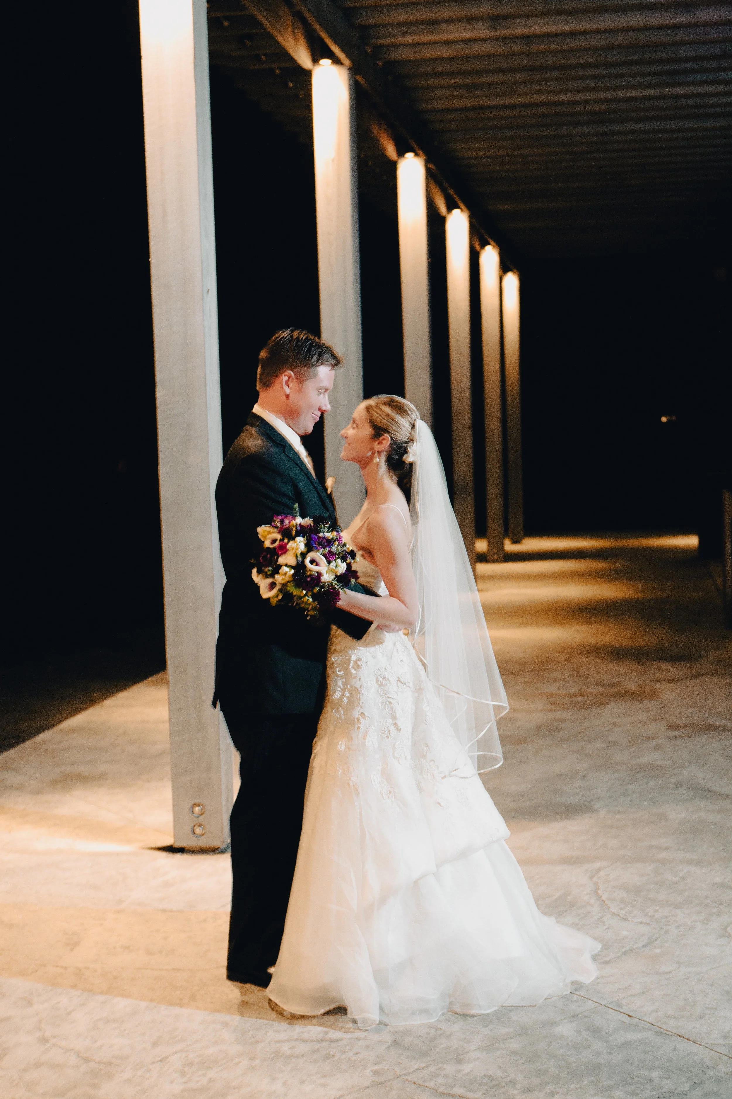 A bride and groom stand close together under a series of tall light fixtures at night, with the bride holding a bouquet of flowers.
