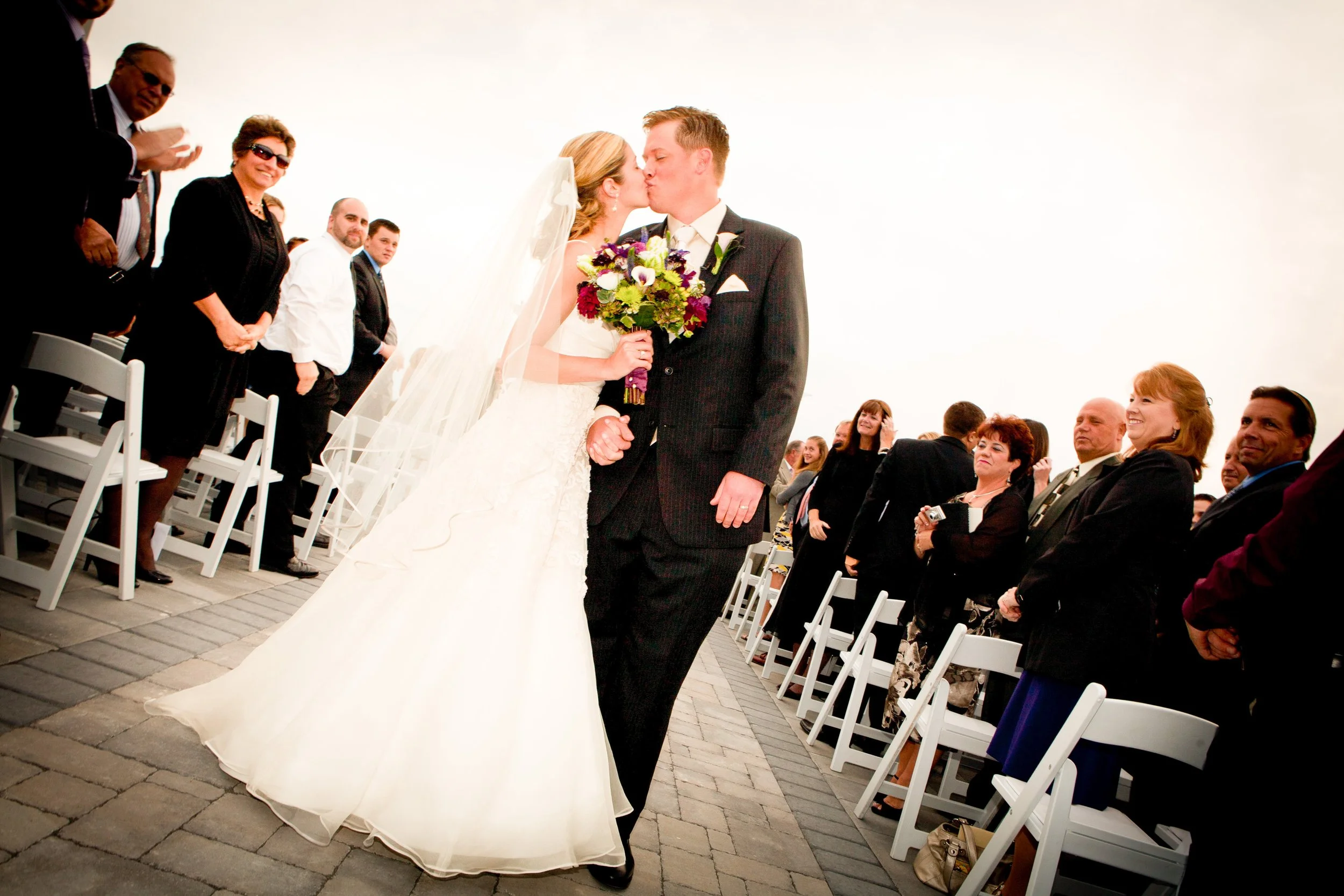 A bride and groom kiss during their wedding ceremony outdoors with guests standing and watching.