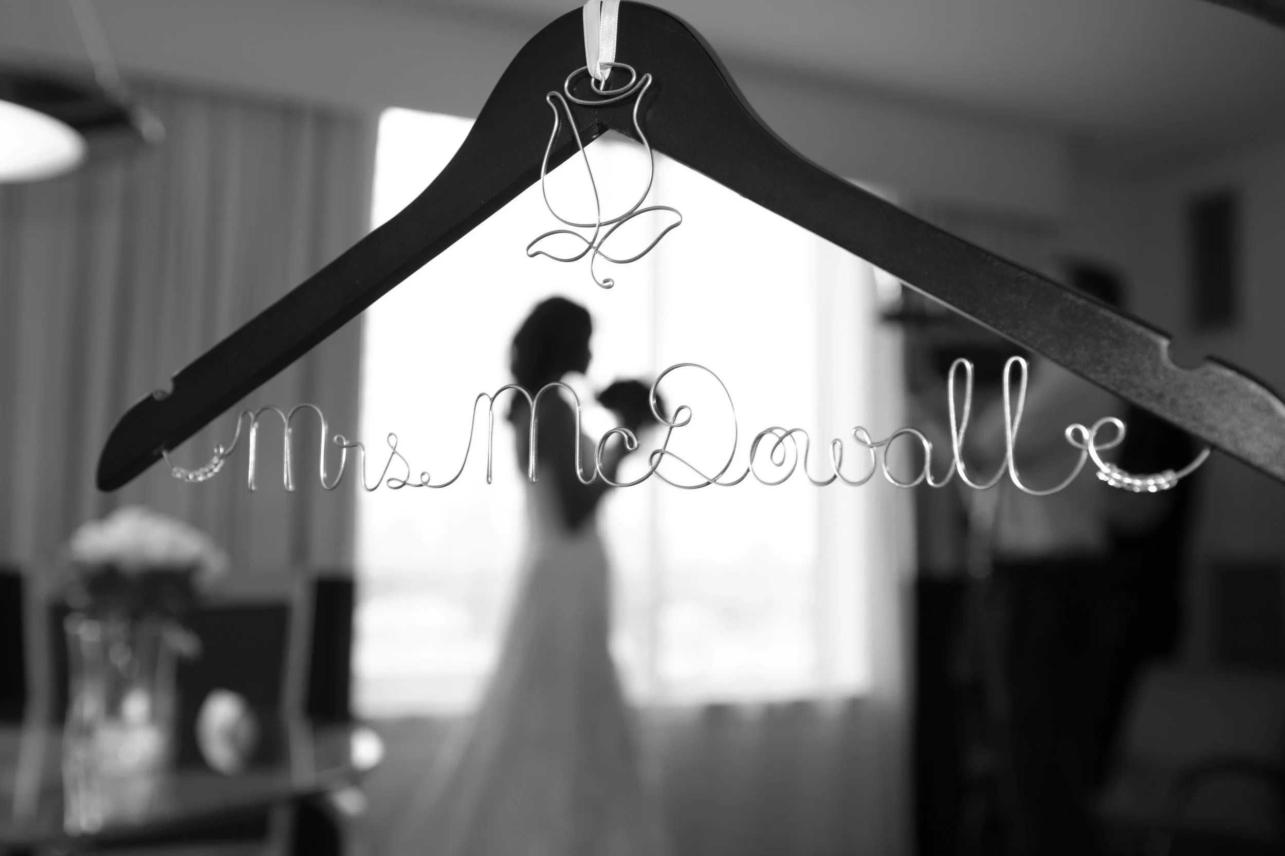 A wire name sign reading "Mrs McDowell" hangs on a black hanger, with a blurred bride in a wedding dress and a person, possibly the groom, in the background