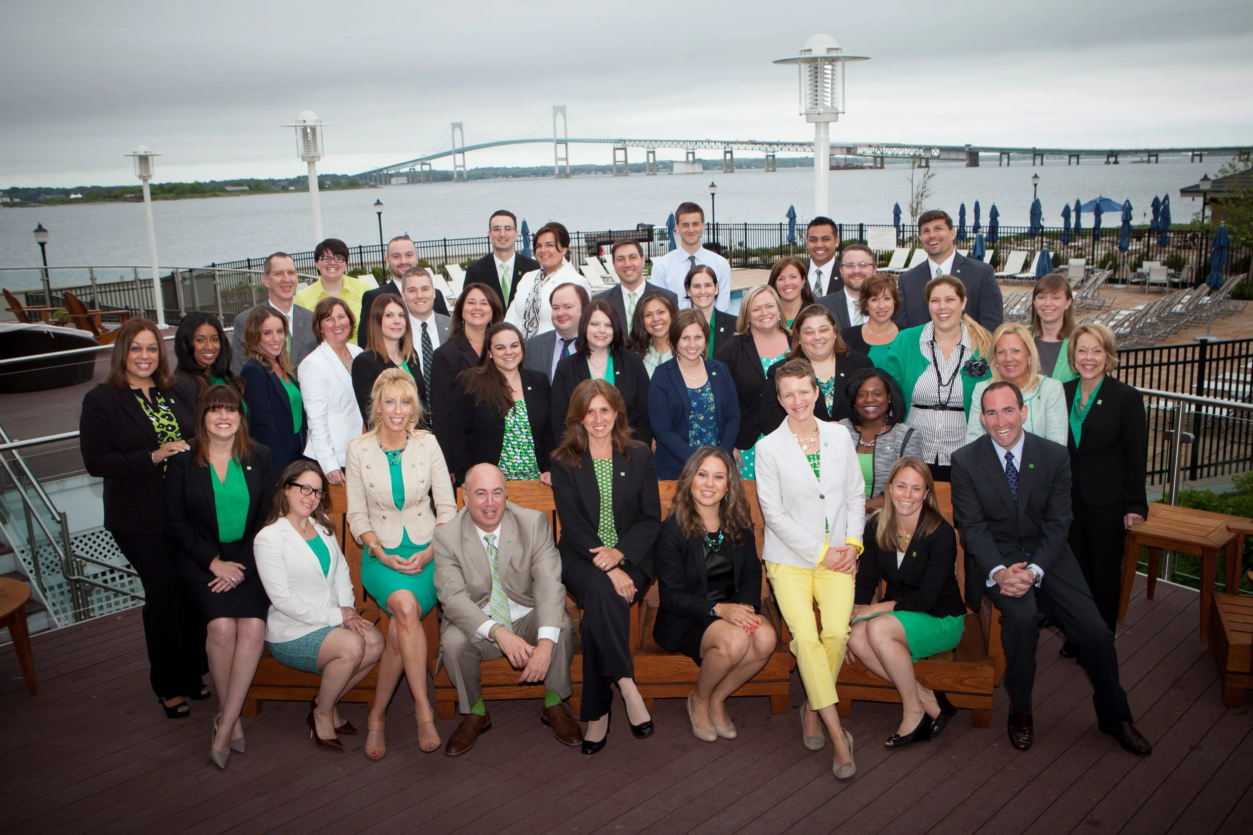 Group of professionally dressed people grouped together on a rooftop deck with water and a bridge in the background.