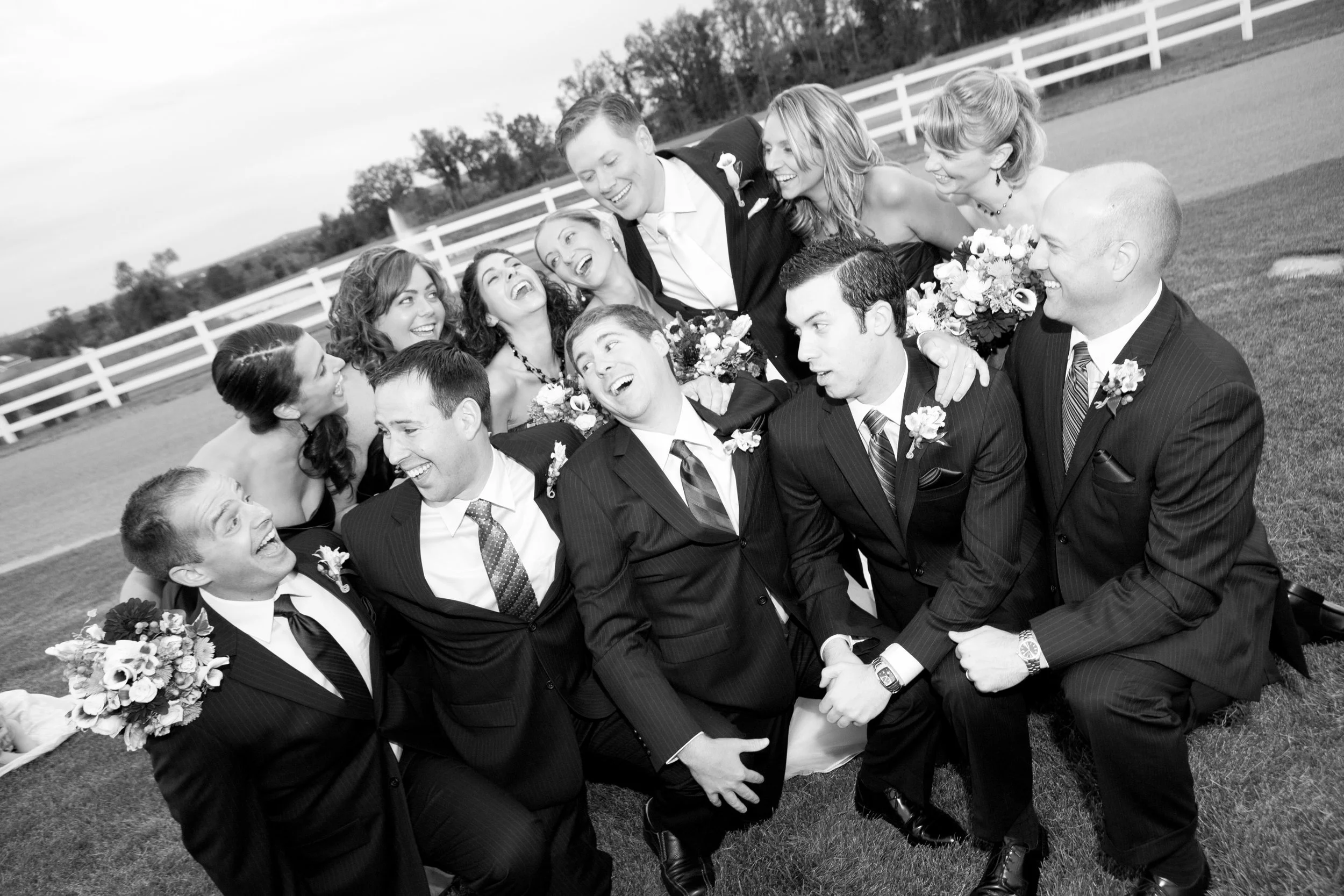 Black and white photo of a wedding party outdoors, with the groom, bride, bridesmaids, and groomsmen, all smiling and laughing, standing on grass near a white fence.